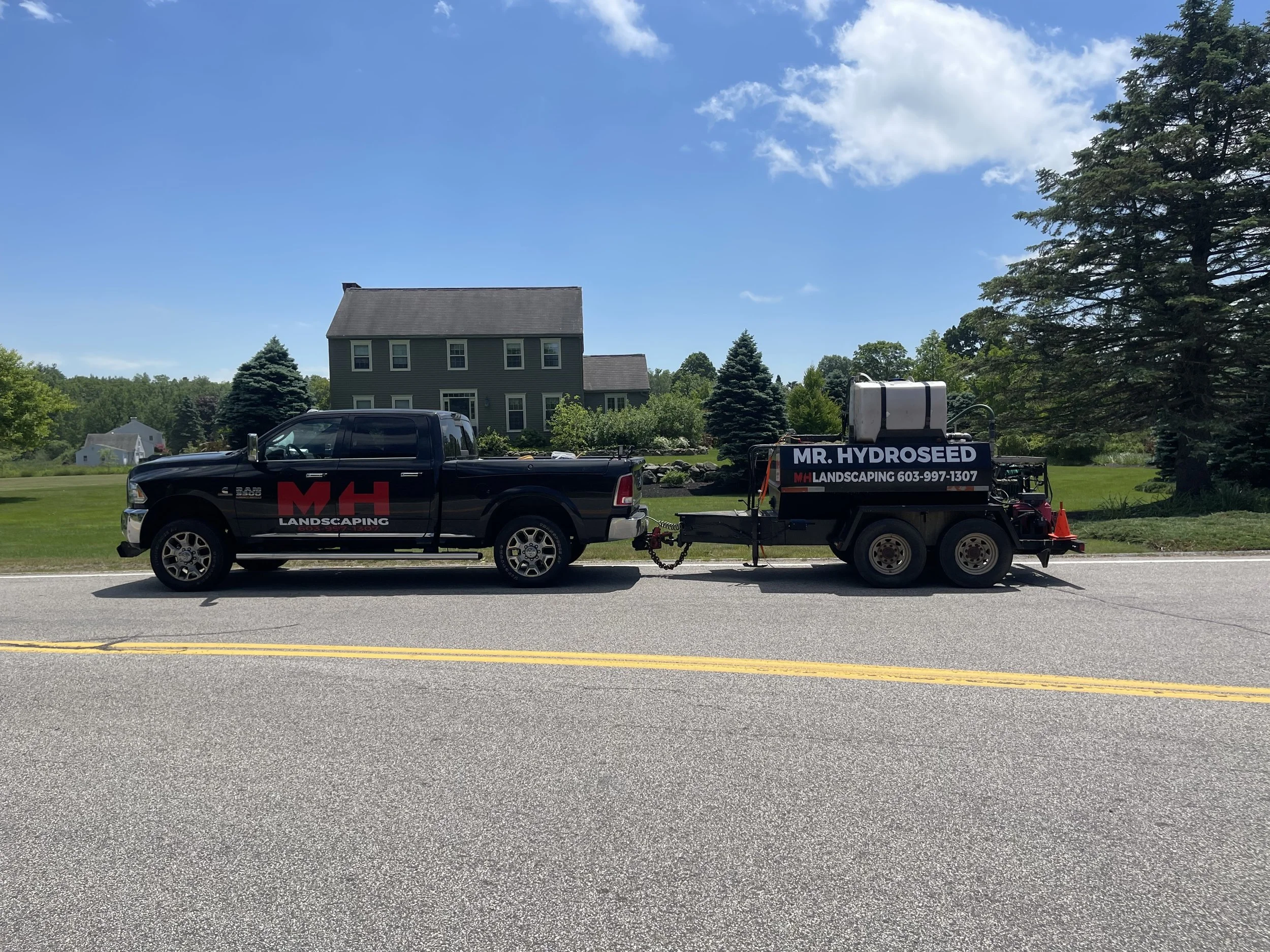 A black truck with the company logo 'MH Landscaping' and phone number, attached to a trailer with landscaping equipment, parked on a road in front of a house with a lawn and trees under a blue sky.