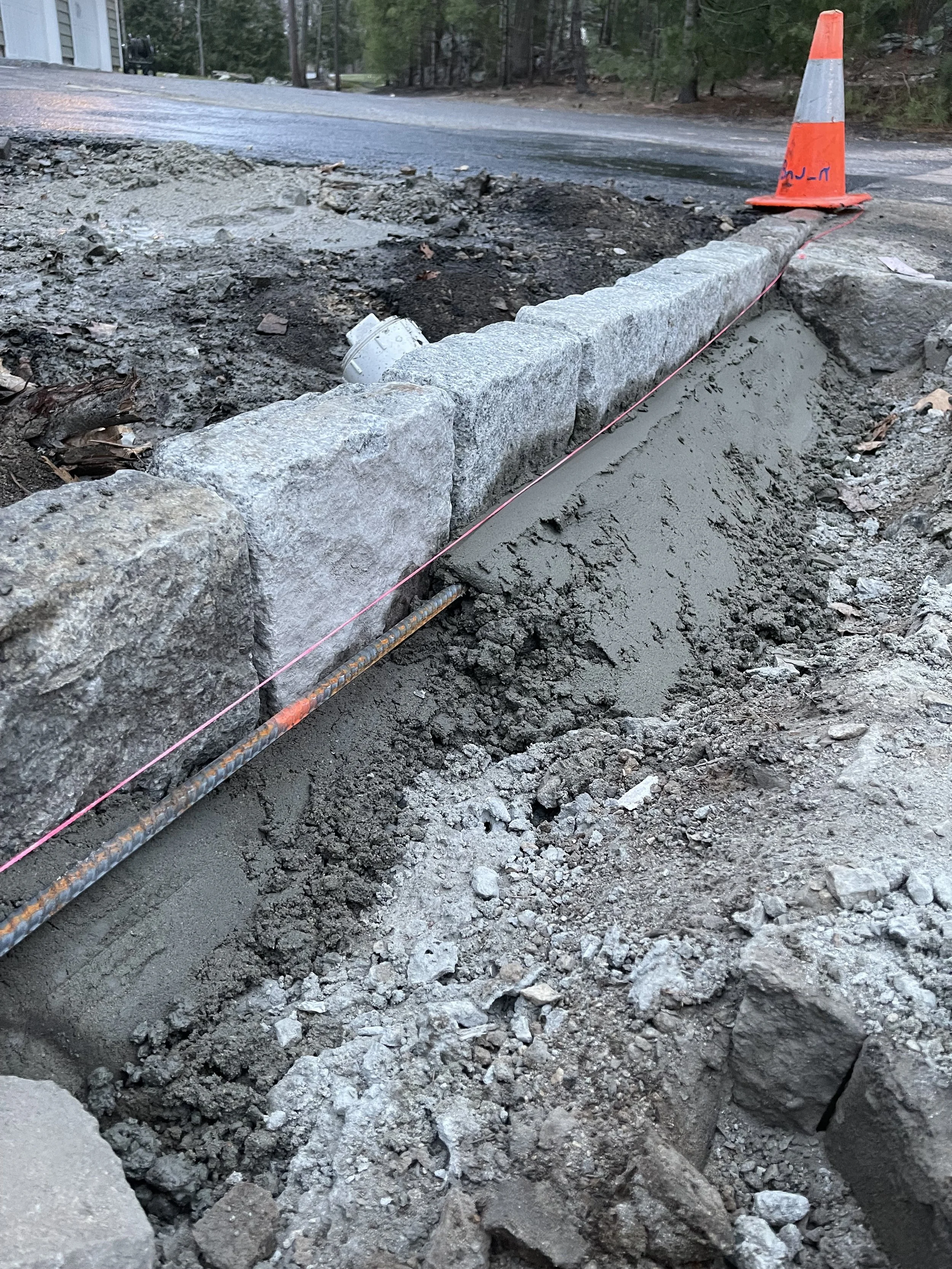 Construction site showing a section of road under repair with a row of gray stone blocks, a cone, and pink construction string.