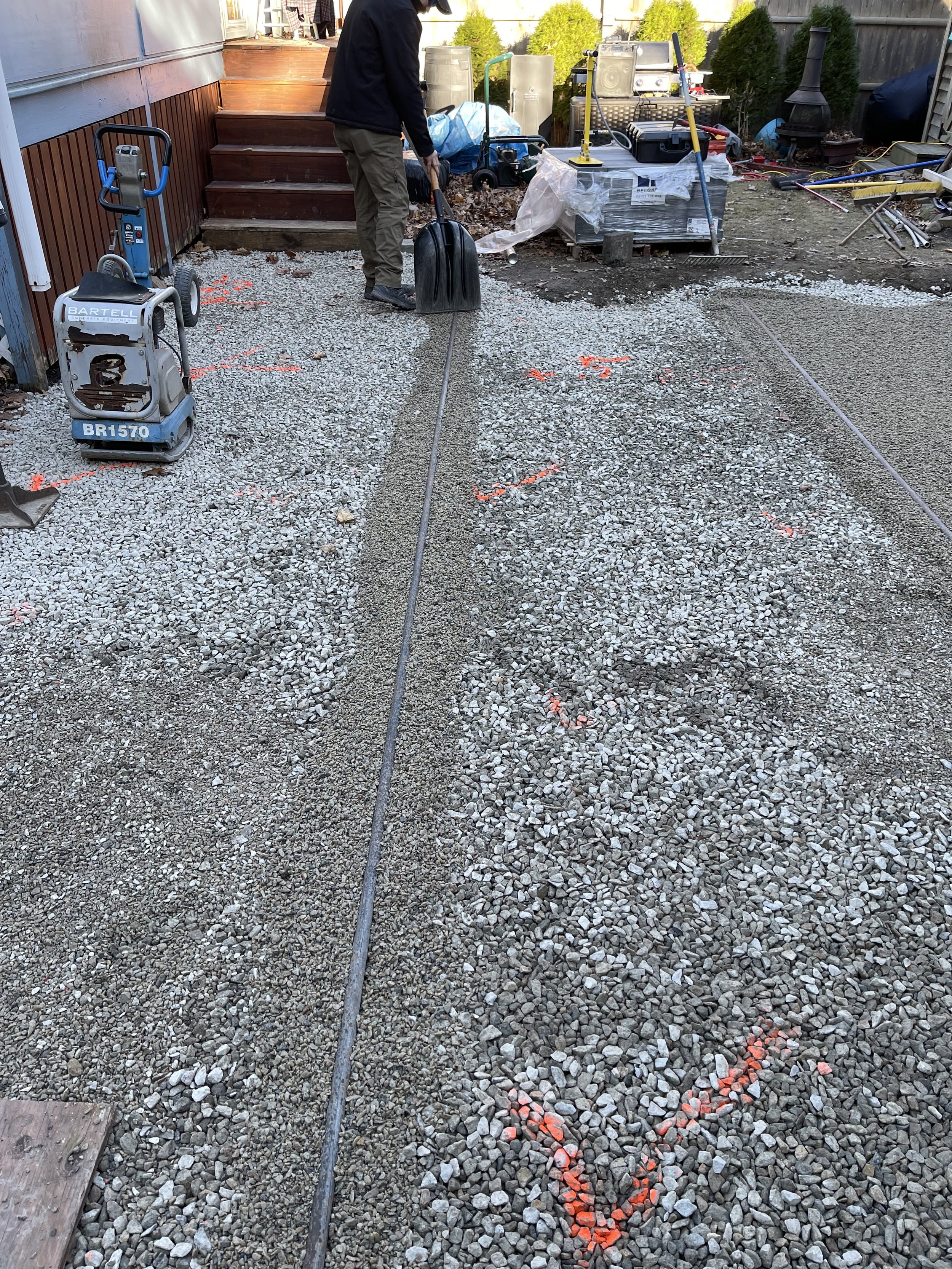 A person working outdoors on a gravel pathway, possibly involved in construction or landscape work. There are tools, equipment, and marked areas on the ground, with a fire pit or chimney in the background and stairs leading to a deck.