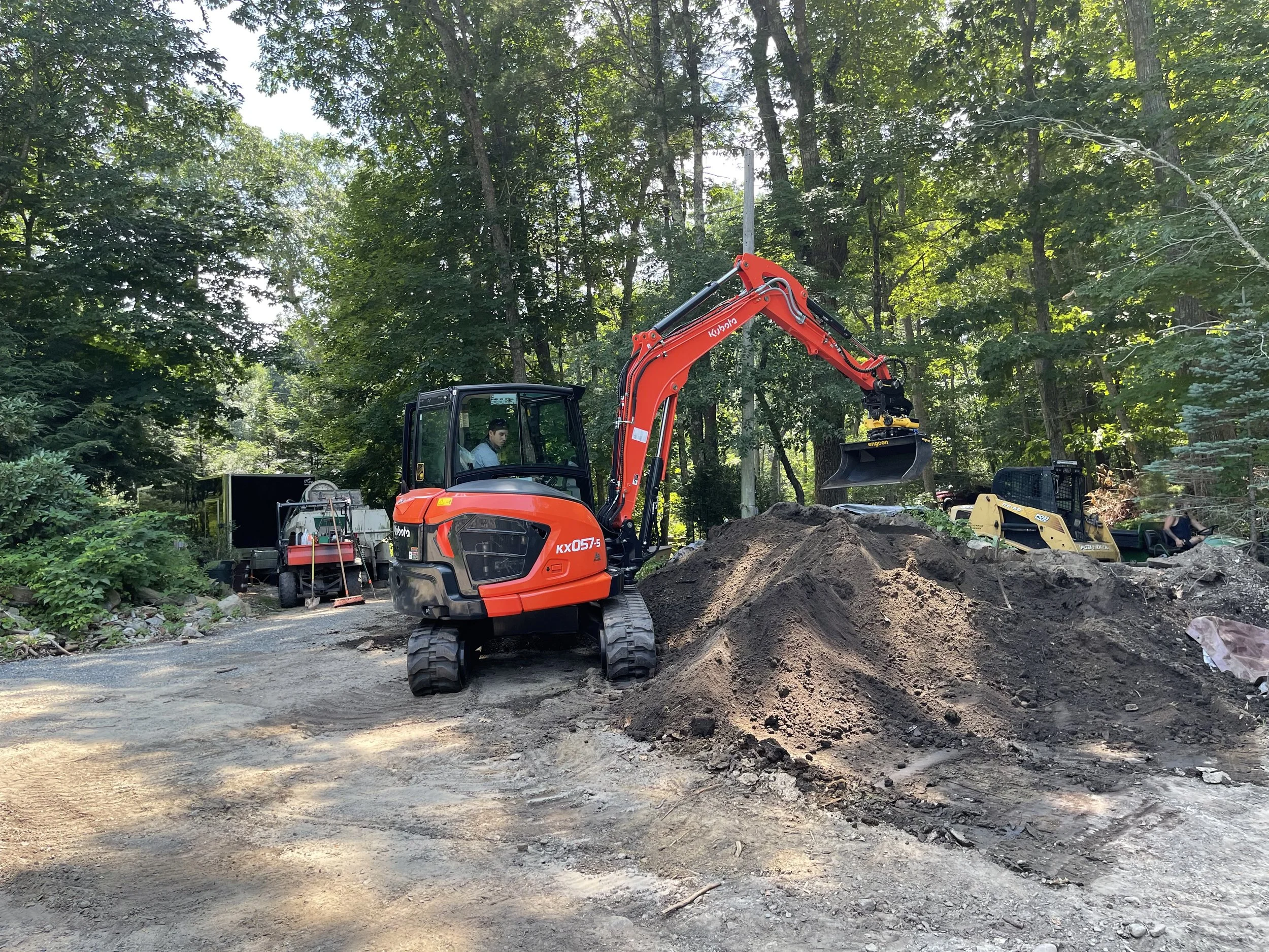A person in a red Kubota excavator moving dirt in a wooded area with trees and other construction equipment visible in the background.