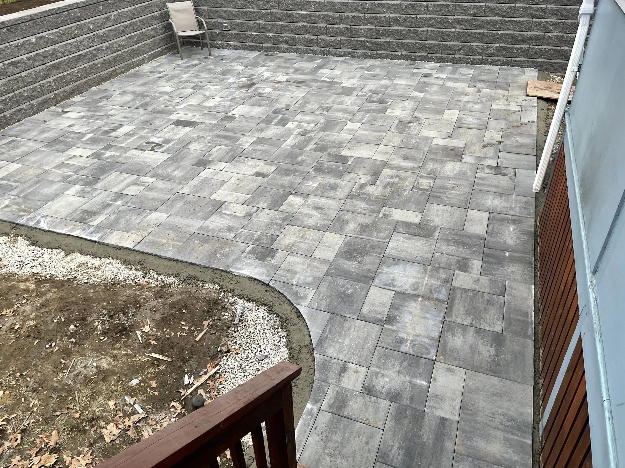 View of a backyard patio with newly installed gray stone pavers. There is a single beige chair near the gray stone wall in the background. The patio edges are bordered by dirt and gravel. On the right side, there is a blue house with a white gutter a