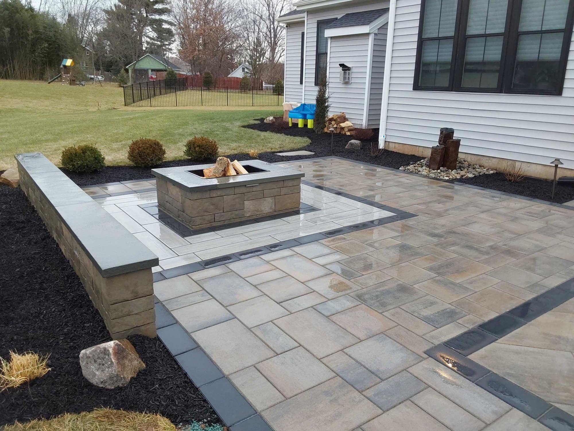 A backyard patio area with beige and gray stone tiles, a fire pit with wood fire inside, decorative rocks, a small water feature, and landscaping with shrubs and mulch next to a house with siding and large windows.