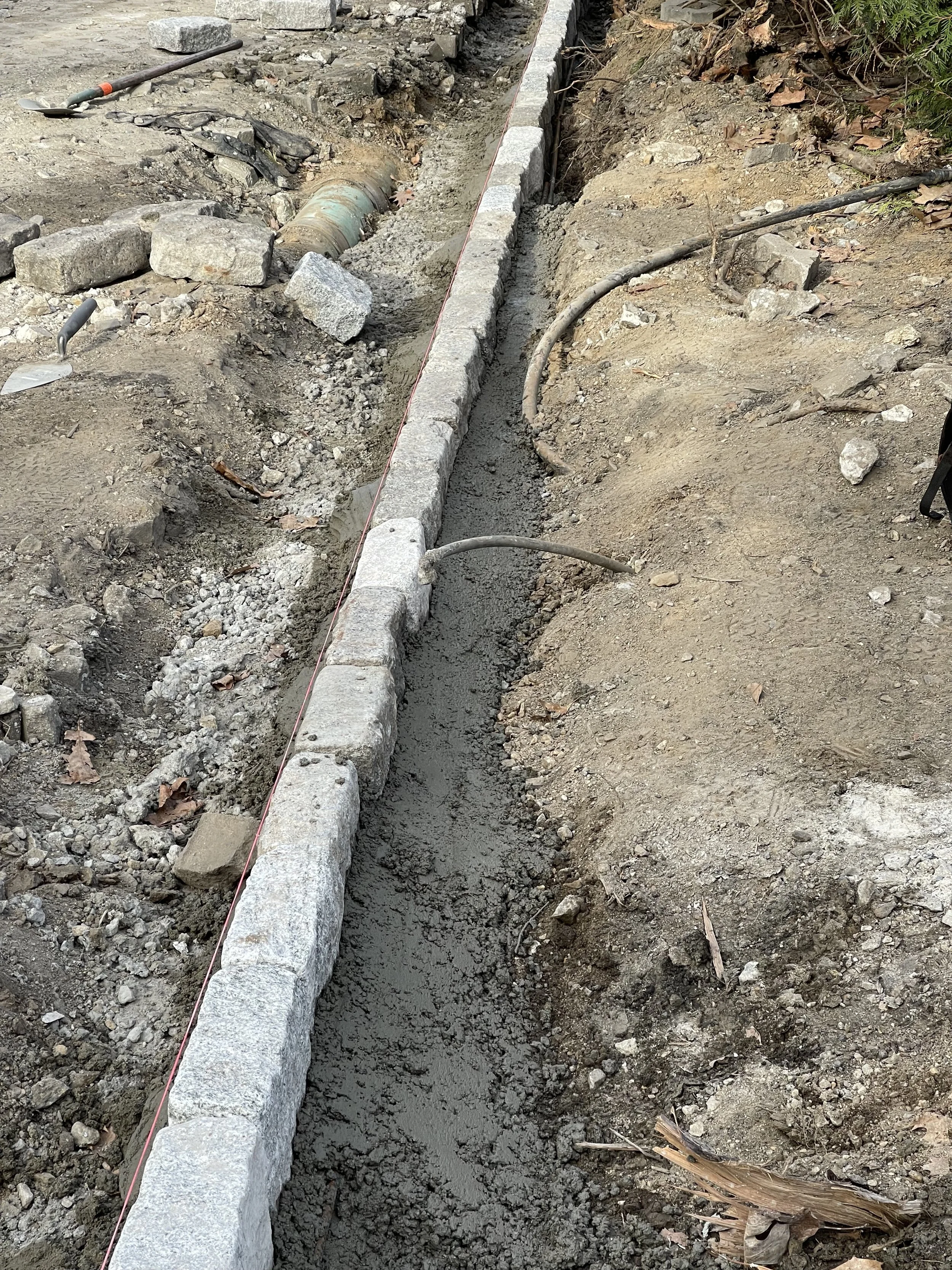 A construction site showing a trench with a row of concrete pavers on the left side, concrete pouring in the middle, gravel, and dirt surrounding the trench, and construction tools like shovels and hoses nearby.