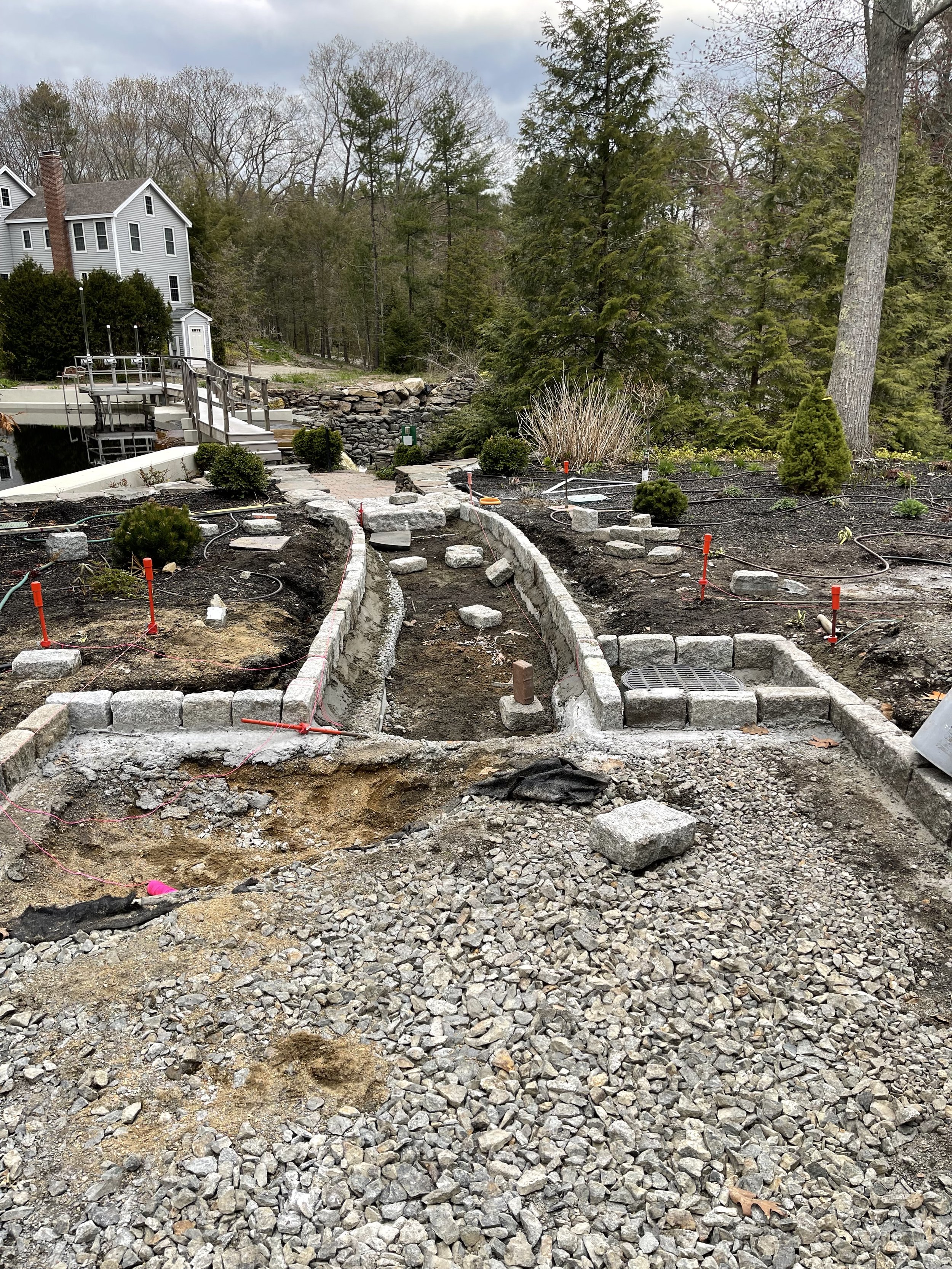 A backyard garden under construction with a curved stone pathway being built, surrounded by gravel, small shrubs, and trees, with a house in the background.