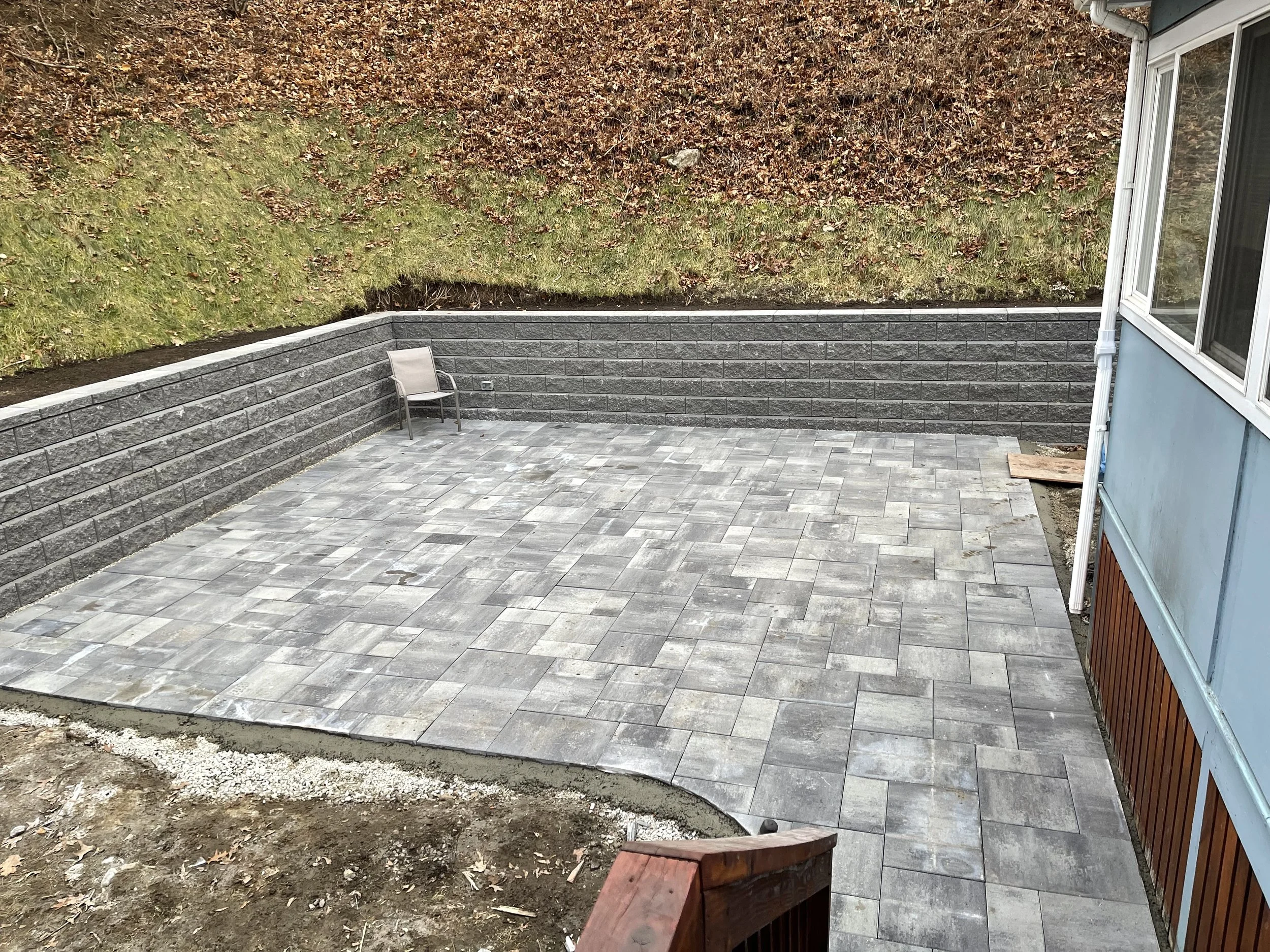 Patio with gray paver stones, wooden bench, gray brick wall, grassy hill with trees and fallen leaves, part of a house with blue siding and white window frames.