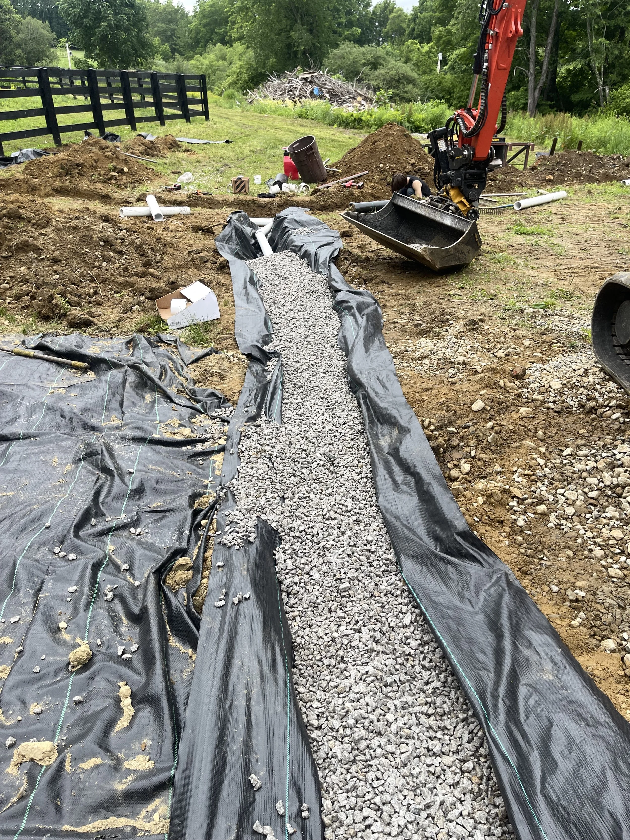 Construction site with black plastic sheeting on ground, gravel being poured into a French trench for drainage, dirt mounds, and construction equipment including a small excavator. There are trees and a fence in the background.