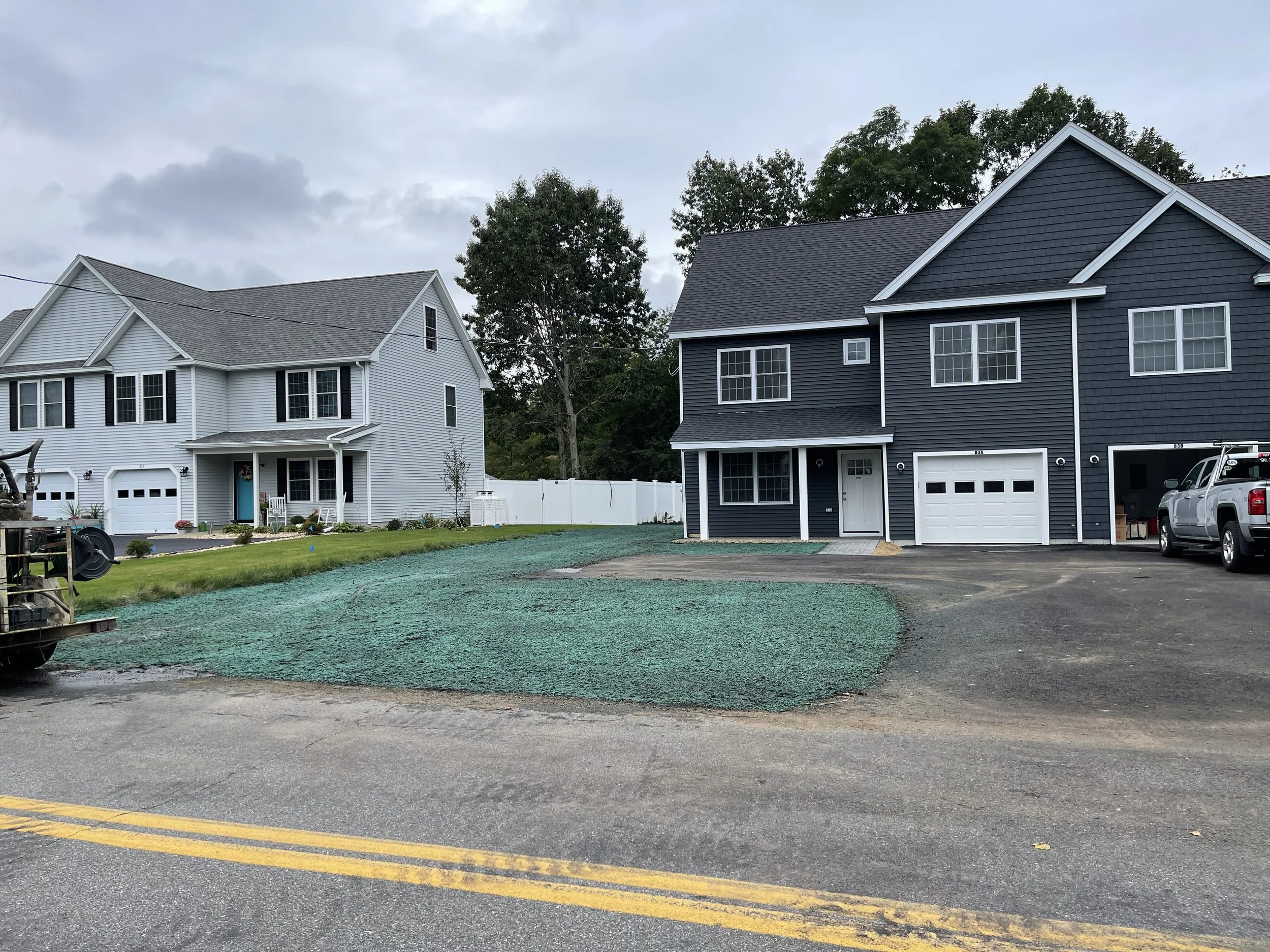 A suburban neighborhood with two houses facing the street. The house on the left is white with black shutters and a blue front door. The house on the right is dark blue with white trim and garage. The sky is overcast, and there is a parking lot with 