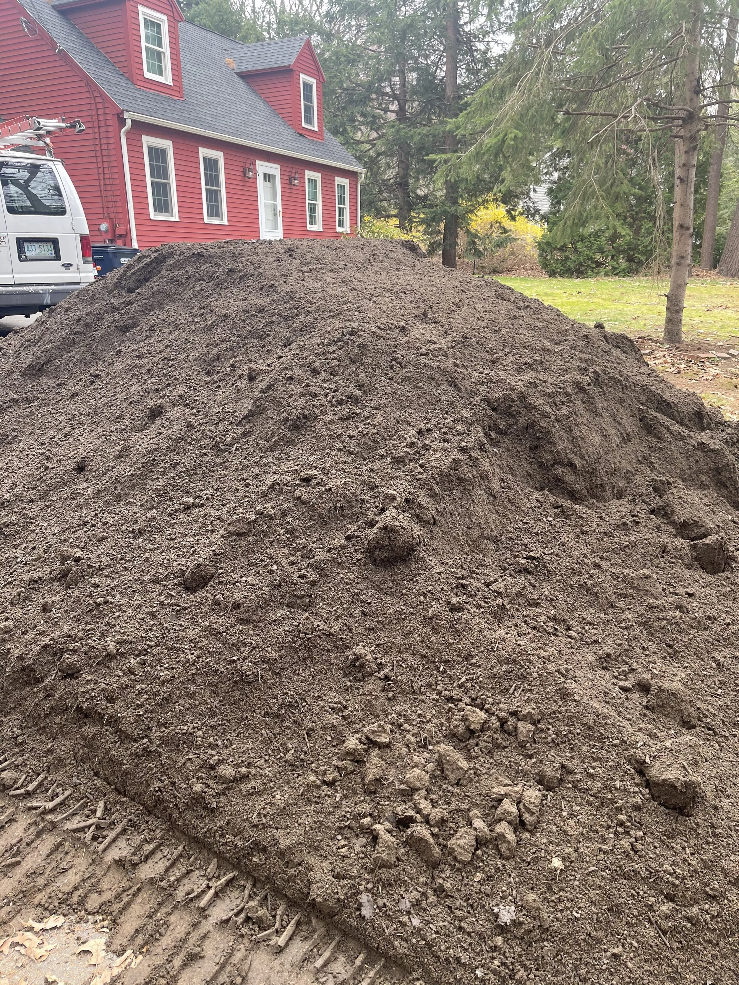 A large pile of dirt in a backyard with a red house in the background, trees, and a white van nearby.