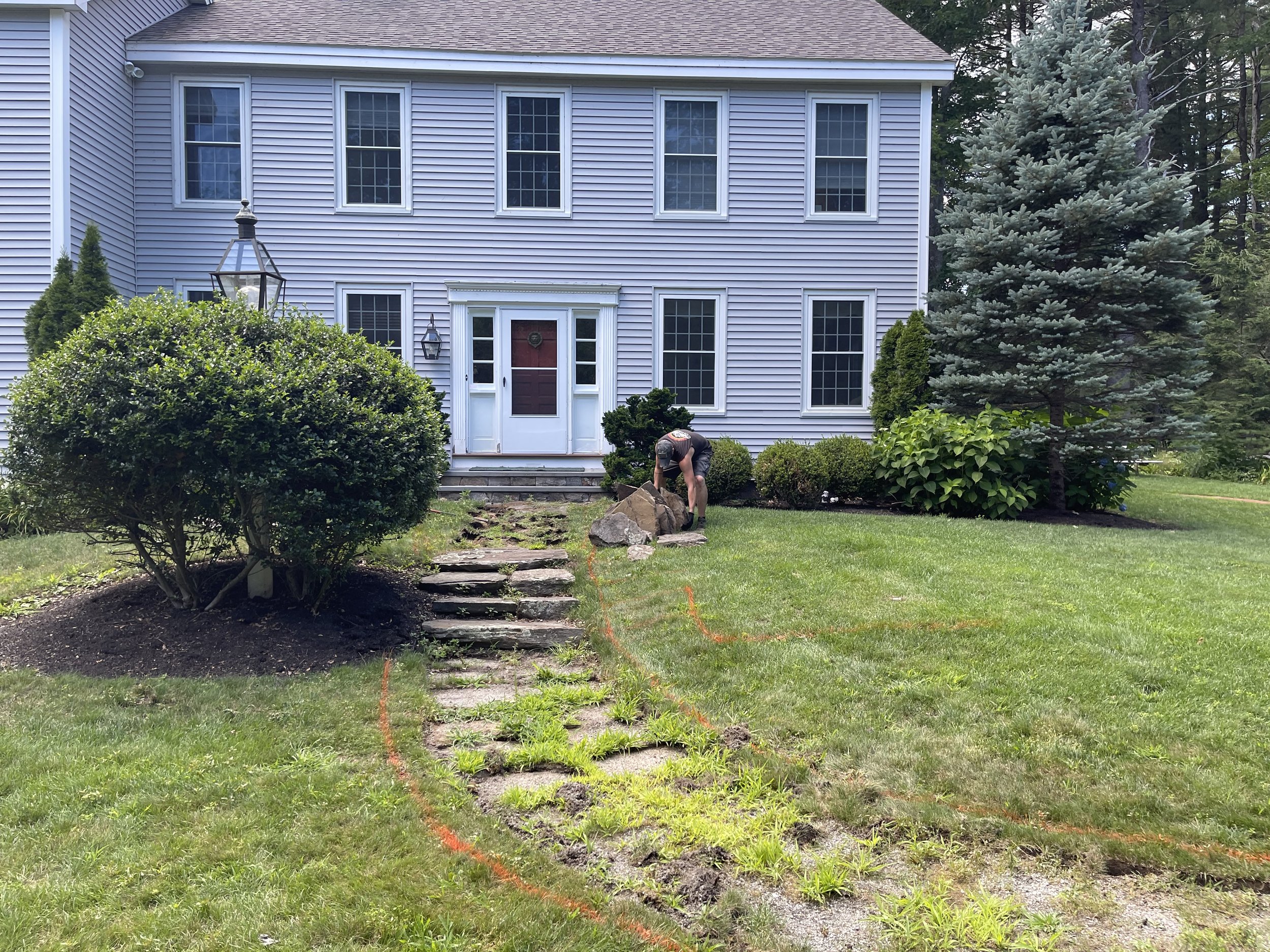 A man working on landscaping in front of a large, light blue house with multiple windows. The yard is being prepared with marked areas for excavation for new brick walkway base gravel , surrounded by bushes and trees.