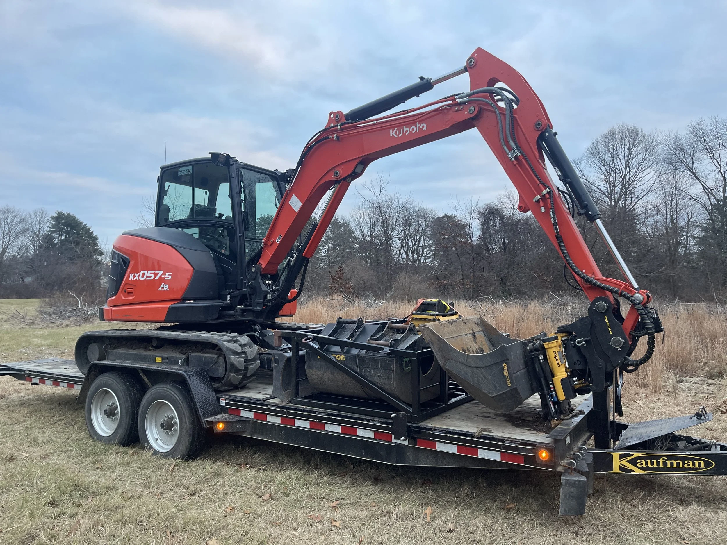 A Kubota KX057-5 mini excavator mounted on a flatbed trailer in an open field with trees and a cloudy sky in the background.