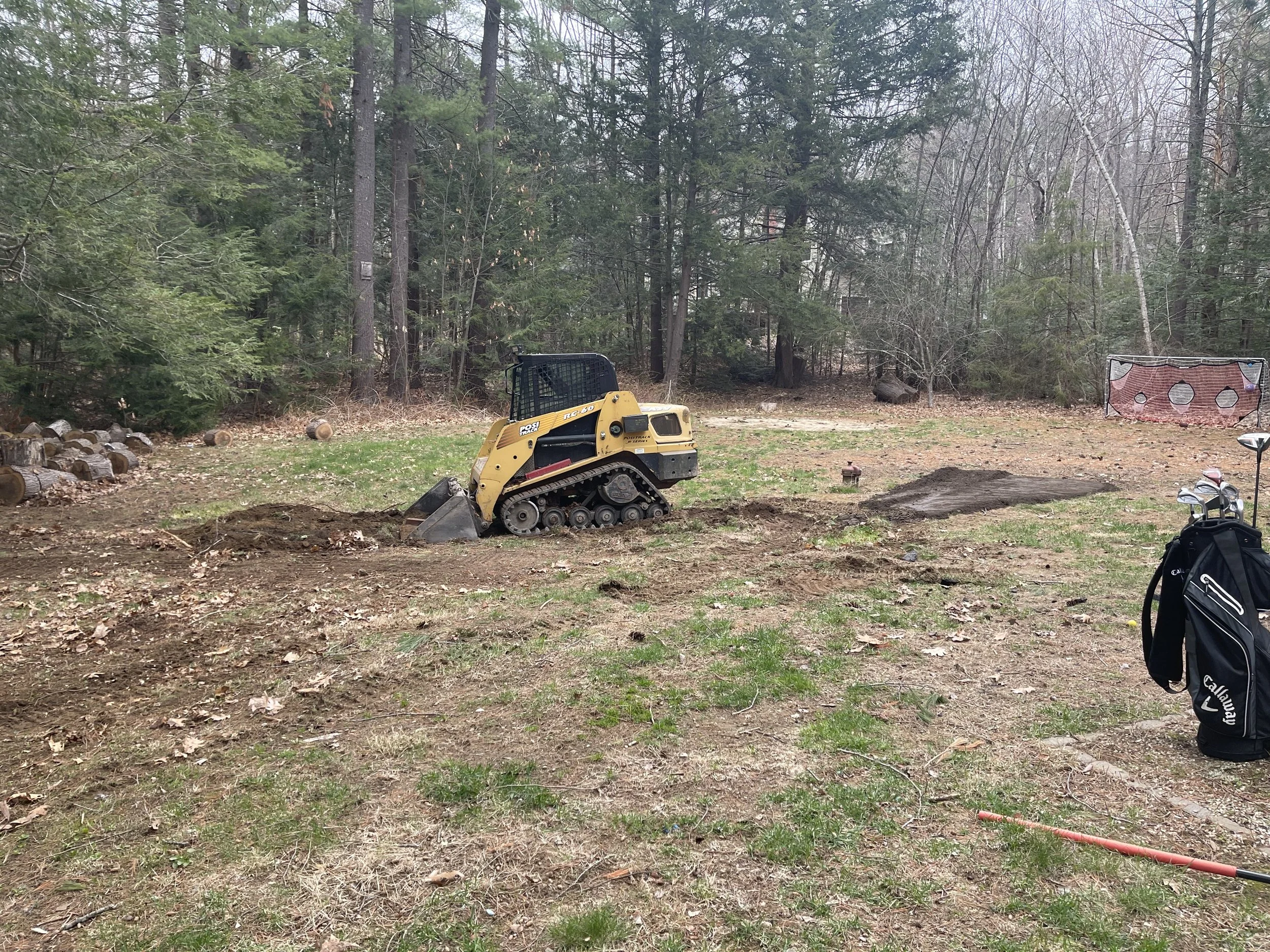 A backyard scene with a small yellow tracked ASV skid steer, a pile of chopped logs, and a sports goal net. The background contains trees with some remaining leaves and some without leaves, indicating a late fall or early spring setting.
