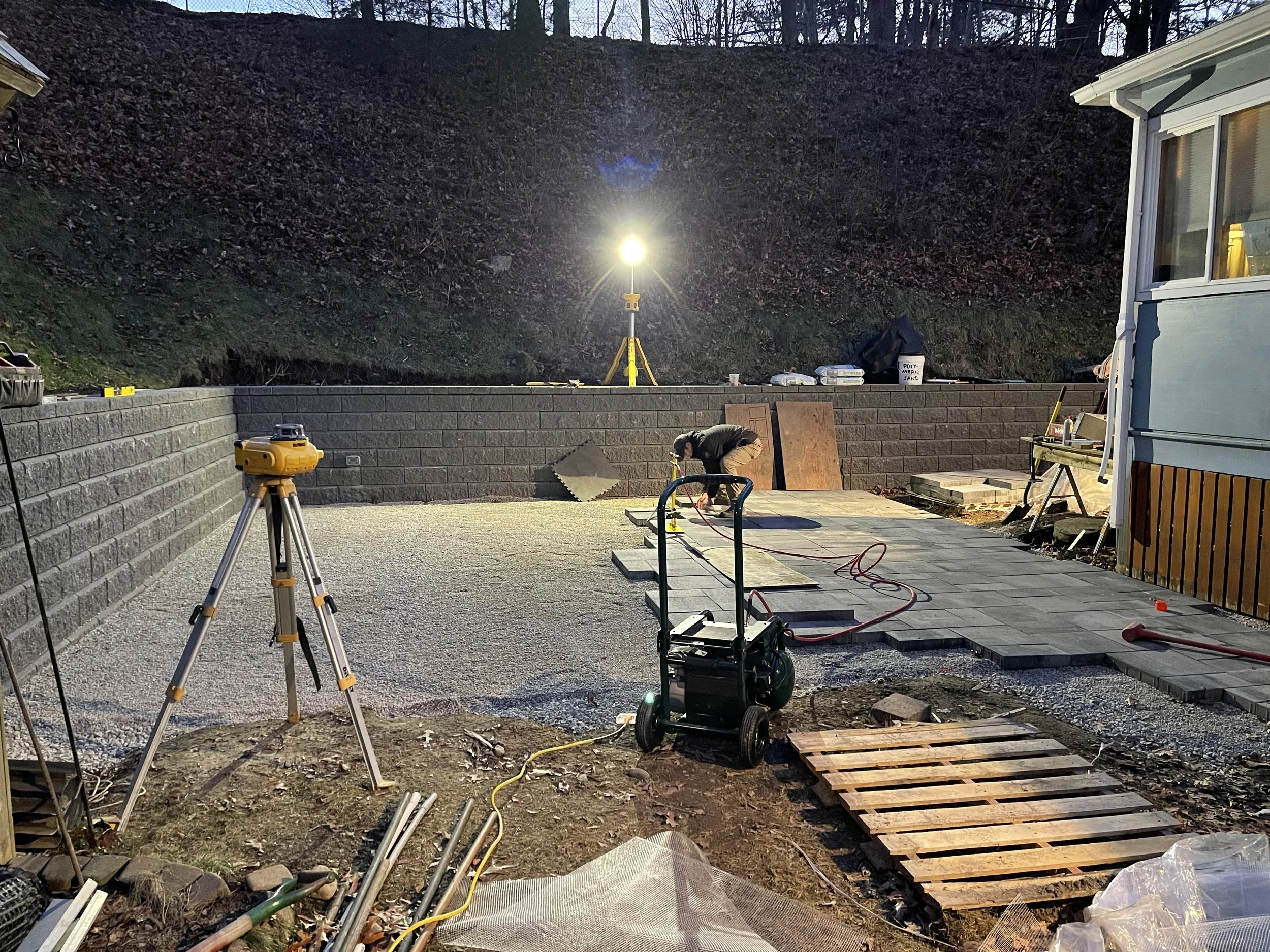 Construction site at dusk with a worker installing paving stones, illuminated by a portable work light, and construction tools and materials scattered around.