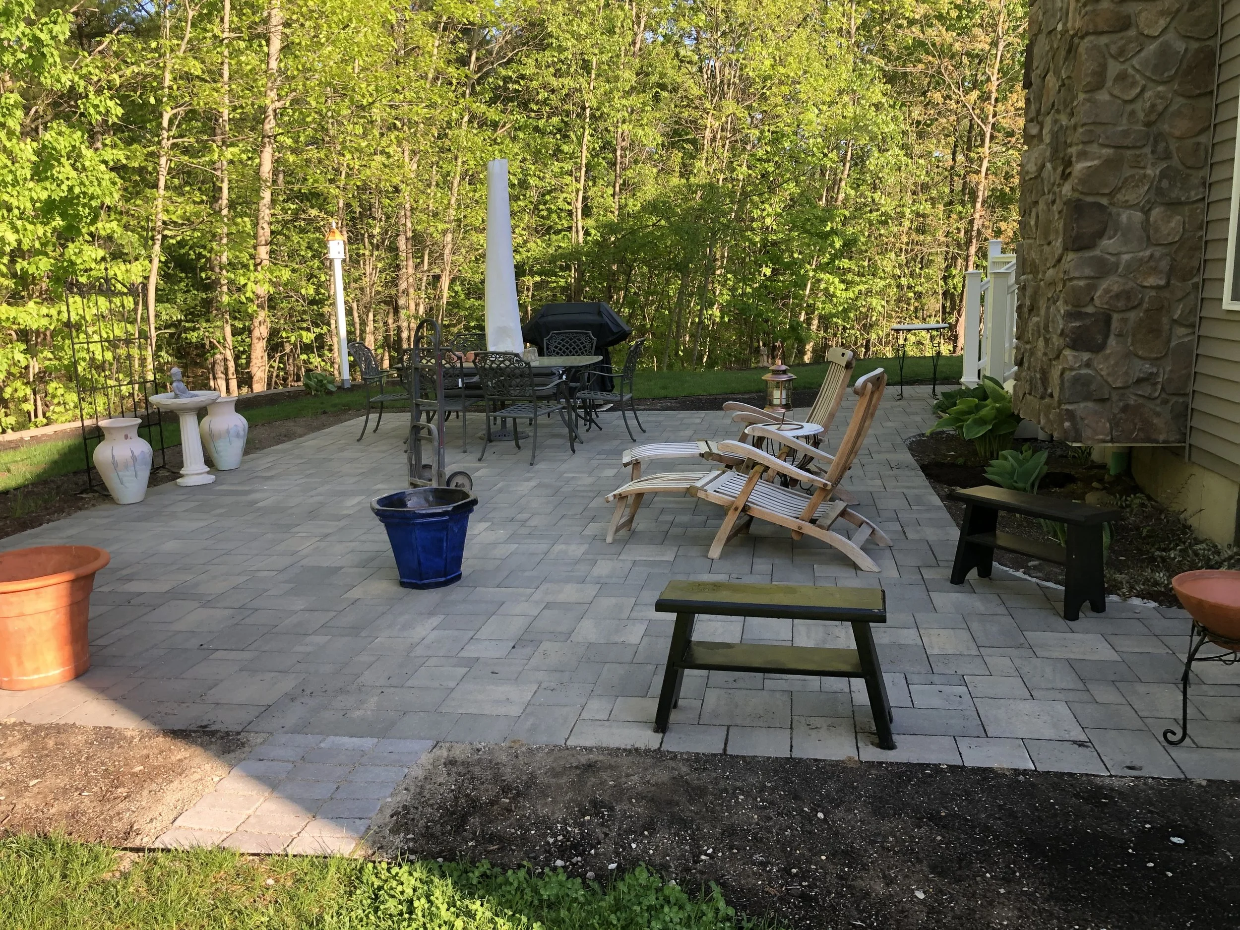 A backyard patio with outdoor furniture including a table with an umbrella and several chairs. There are decorative vases, potted plants, and a birdhouse visible. The patio is surrounded by trees and greenery, with some parts of the paving stones and