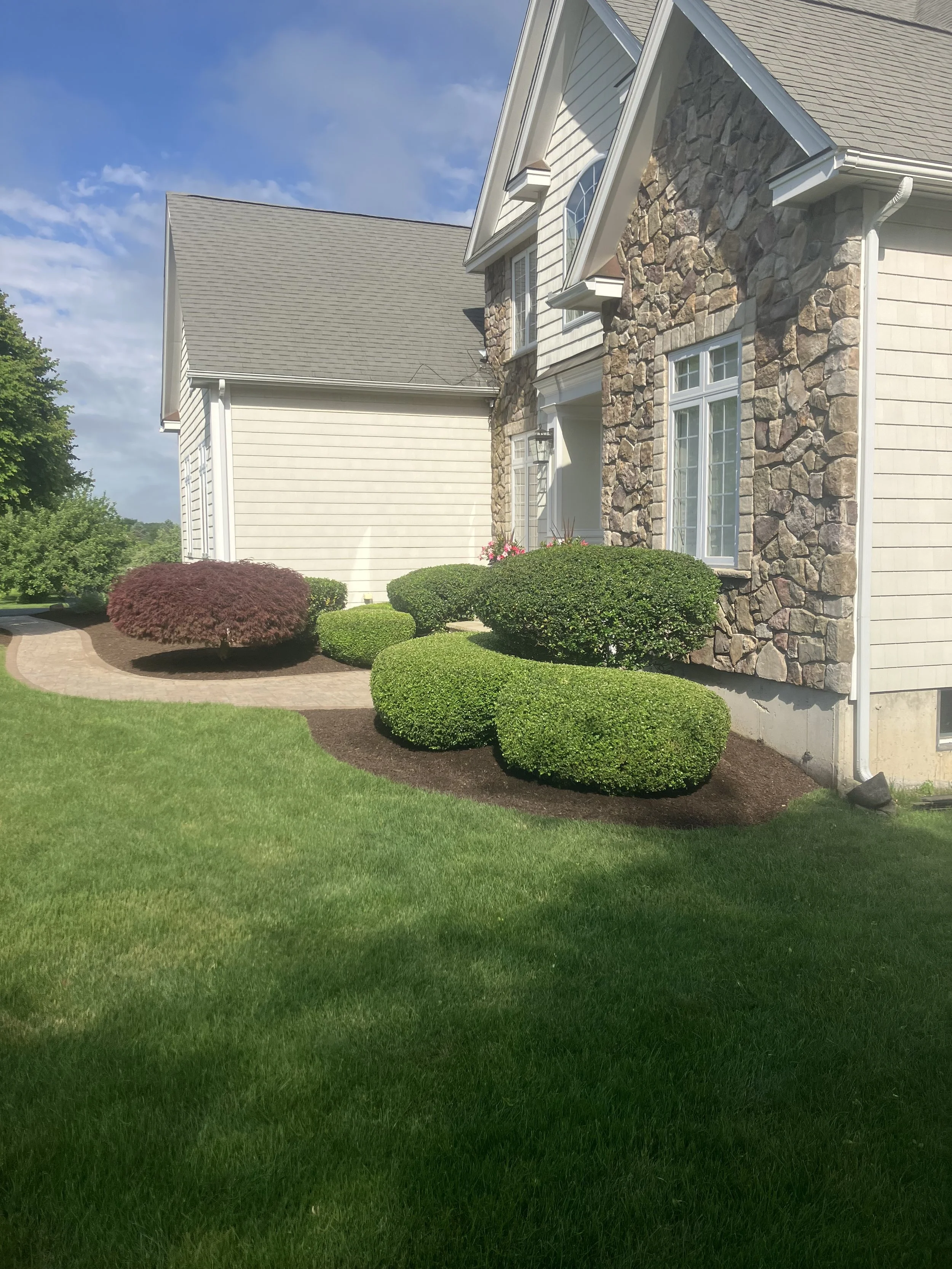 Front yard with neatly manicured green lawn, shrubs, and a two-story house with stone and siding exterior under a partly cloudy sky.