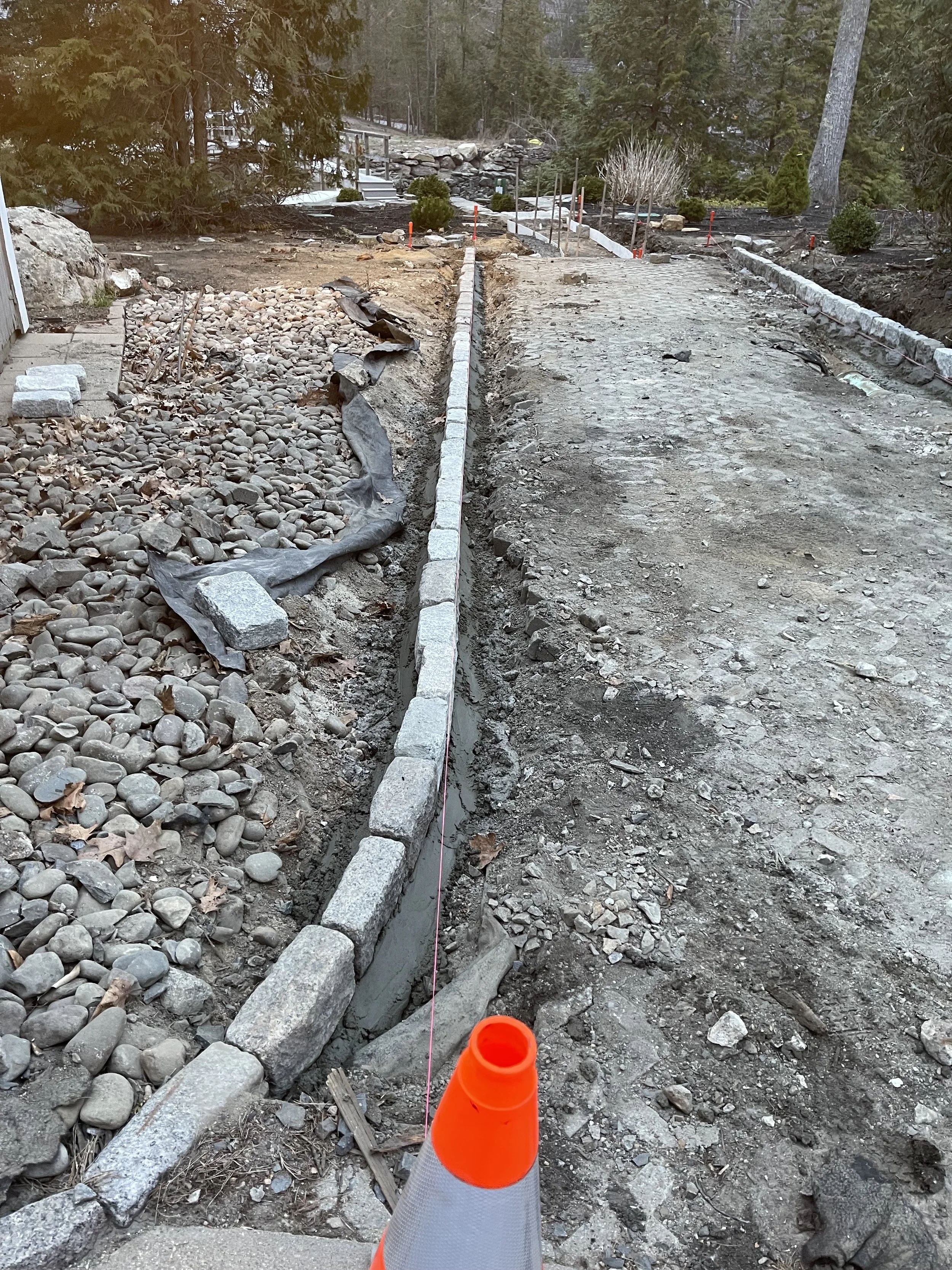 A construction site showing a partially built sidewalk or pathway, with a string line for alignment, orange safety cone in the foreground, and trees in the background.