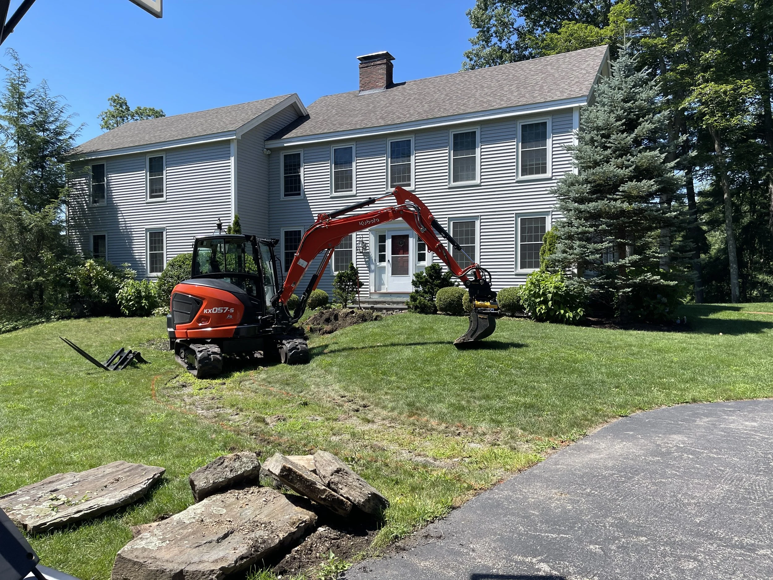 A house with white siding and multiple windows is under renovation, with a small red excavator working on the yard, surrounded by green trees and a clear blue sky.