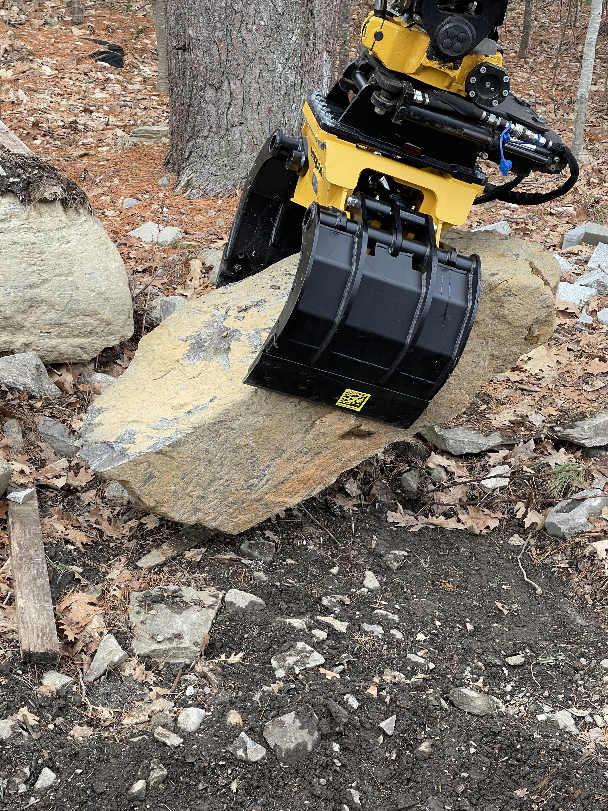 A yellow and black hydraulic excavator digging soil and rocks in a wooded area, with a tree and fallen leaves in the background.