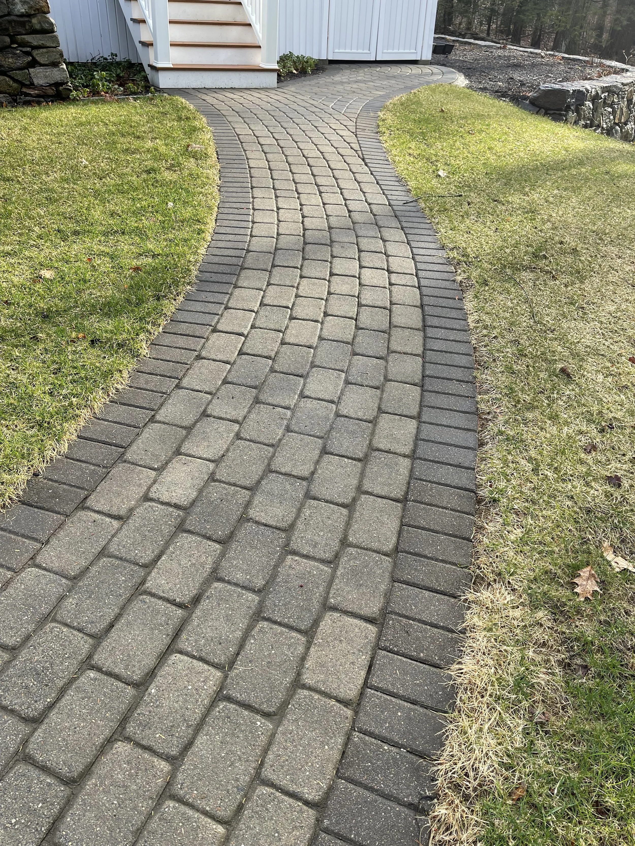A paver walkway made of rectangular Techobloc pavers in a curved pattern, bordered by grass on both sides, leading to stairs beside a house with white siding and a stone retaining wall on the right.