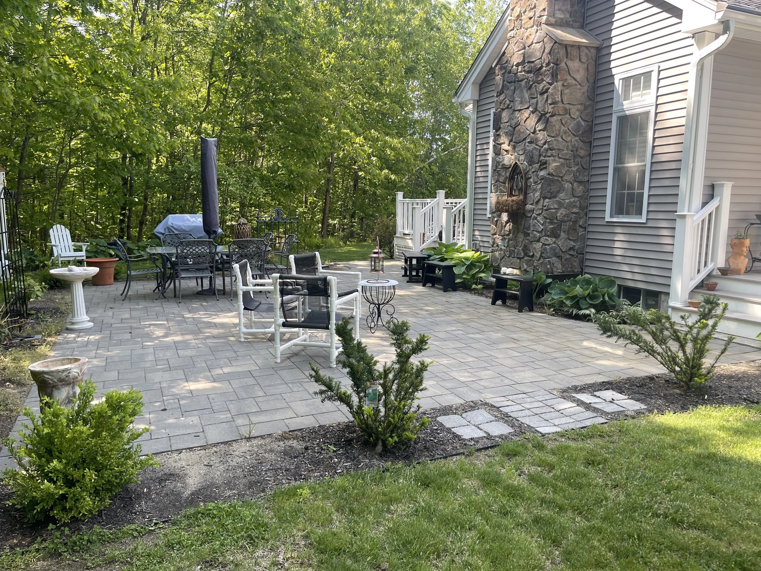 A backyard patio with outdoor furniture, surrounded by trees and plants, with a house featuring a stone chimney on the right.