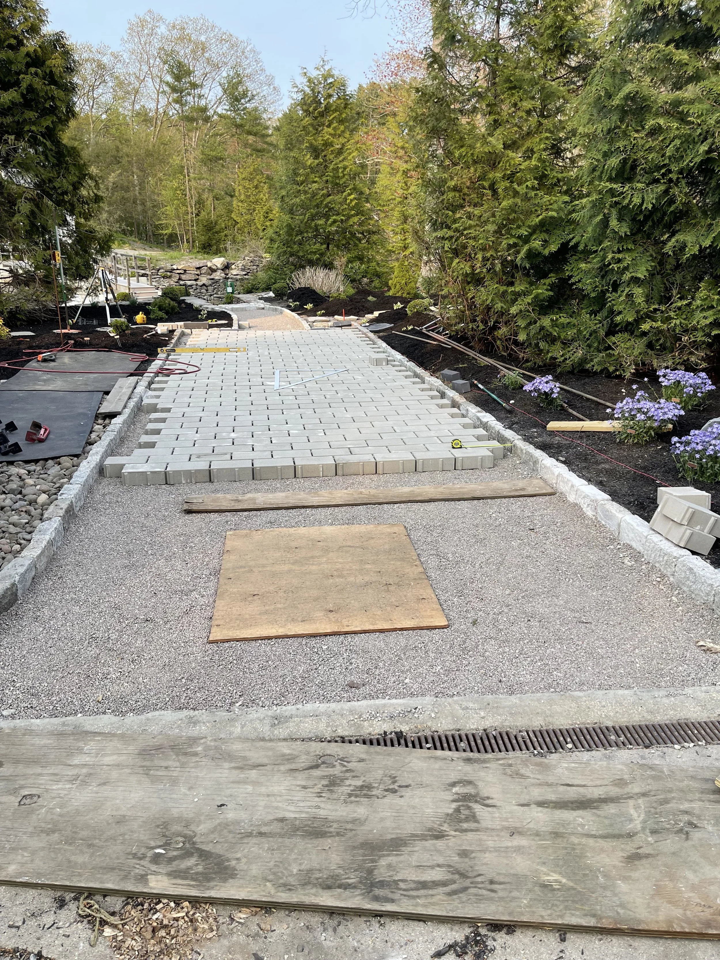 A pathway with newly laid bricks, surrounded by greenery and flowers, with tools and construction materials on the sides, under a clear sky.