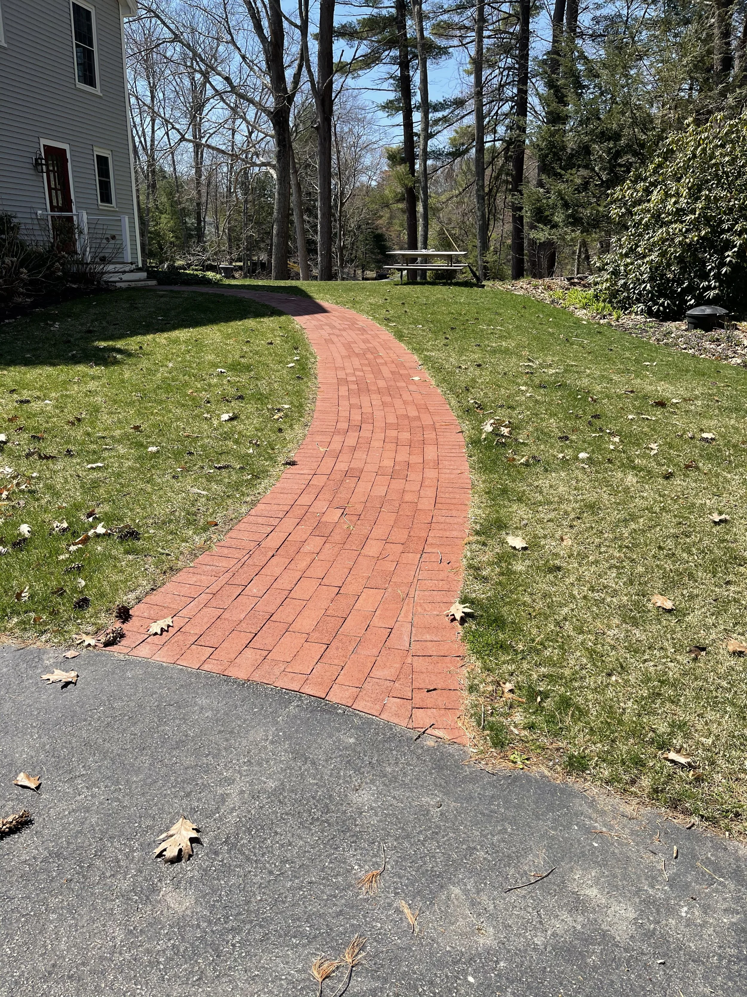 A brick pathway curves through a green lawn with scattered fallen leaves. To the left is a house with beige siding and white trim. In the background, there are tall trees with some leaves and bare branches, and a picnic table is visible in the distan