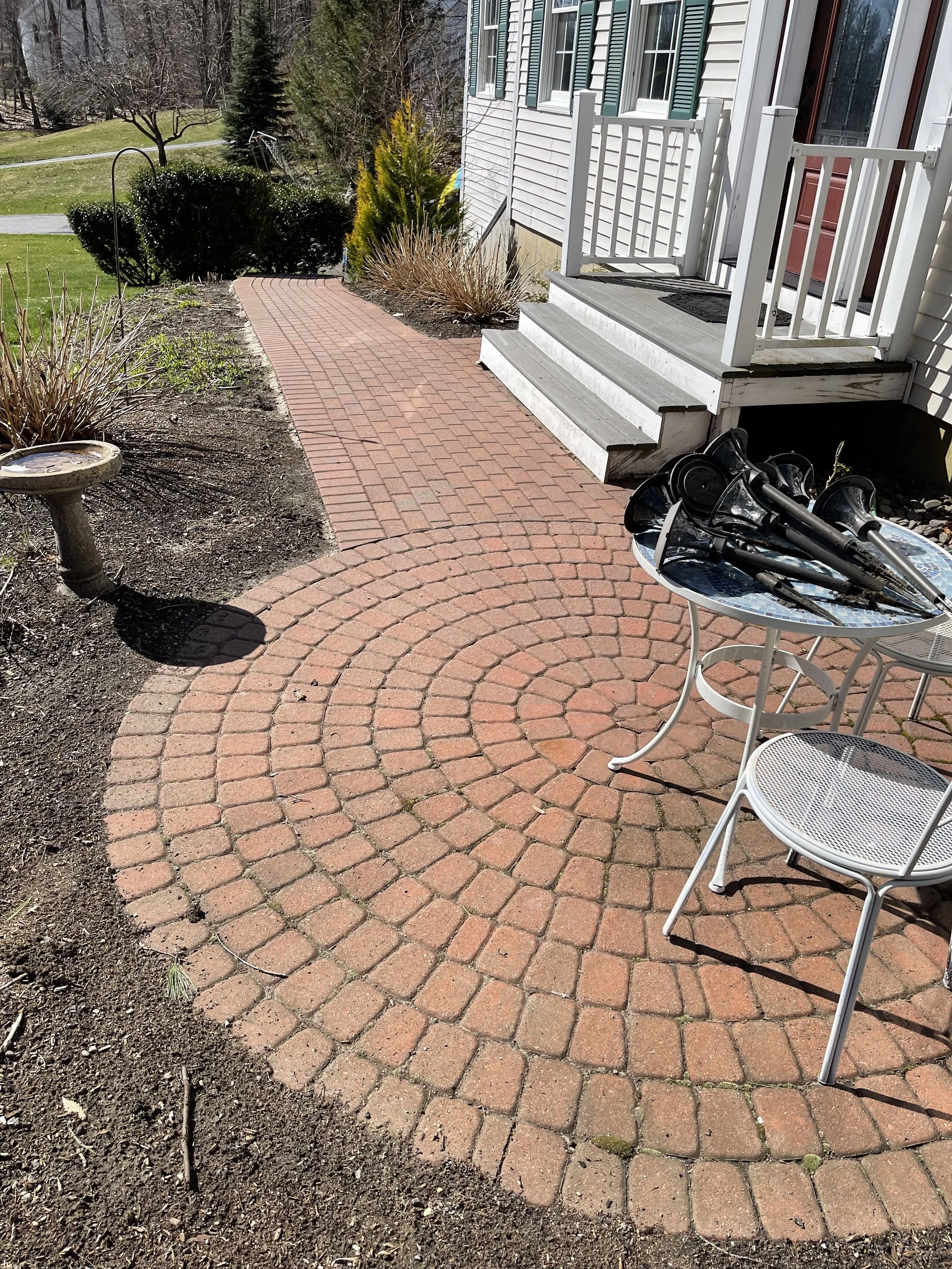 A brick patio with stairs leading up to a house door. There are outdoor tables and chairs, gardening tools, bushes, and a bird bath in the yard.