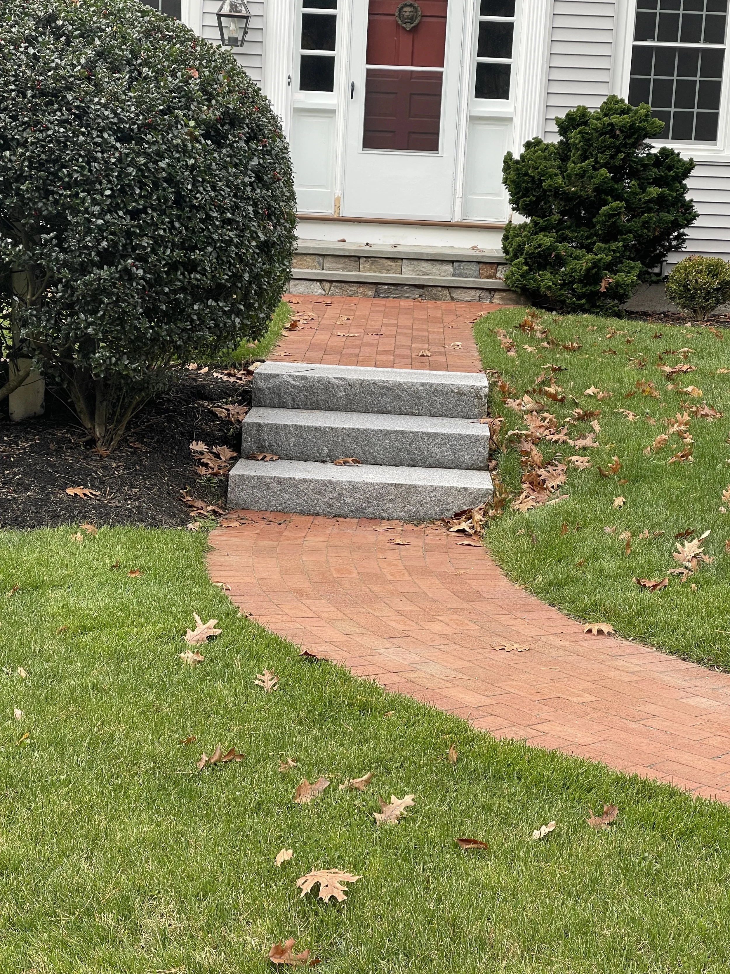A front yard with green grass, fallen autumn leaves, a red brick pathway leading to a house with a porch, surrounded by bushes, and a white door with glass panels.