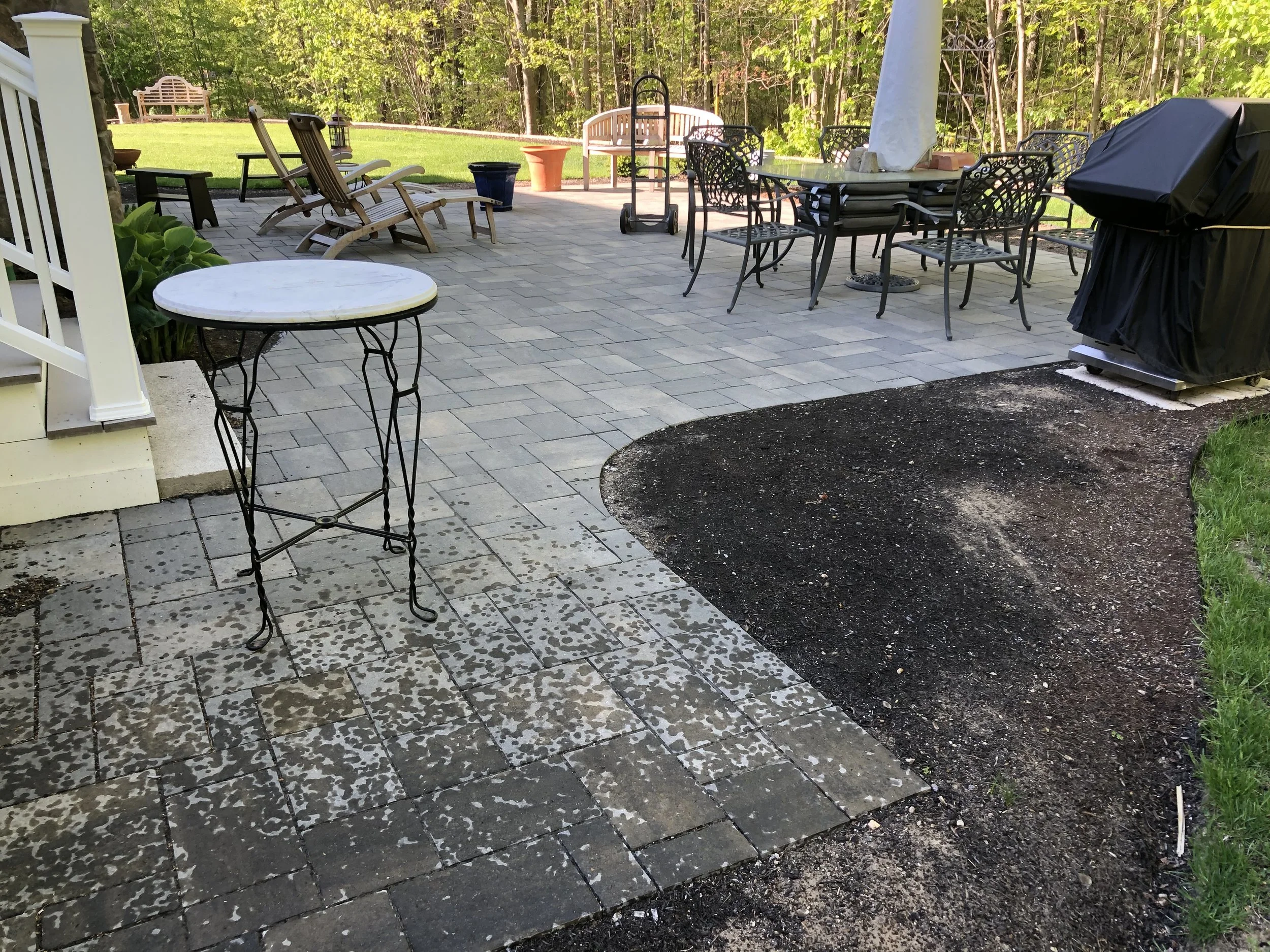 Backyard patio area with patio furniture, including chairs, table, and a covered grill on the right. A grassy yard with trees and a bench in the background.