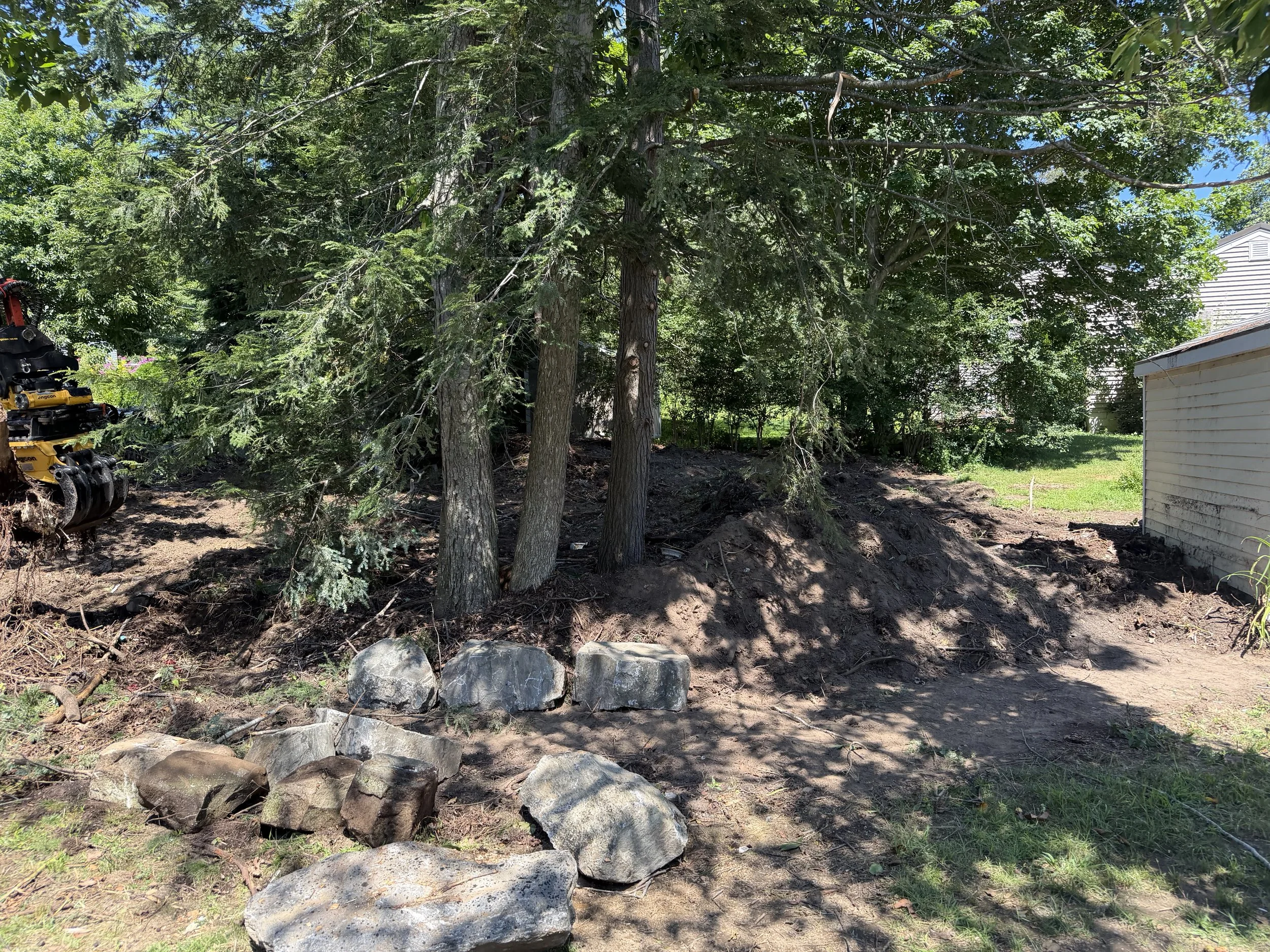 A backyard with tall trees, a dirt area with a row of large rocks, a neighboring house on the right, and some construction equipment partially visible on the left.