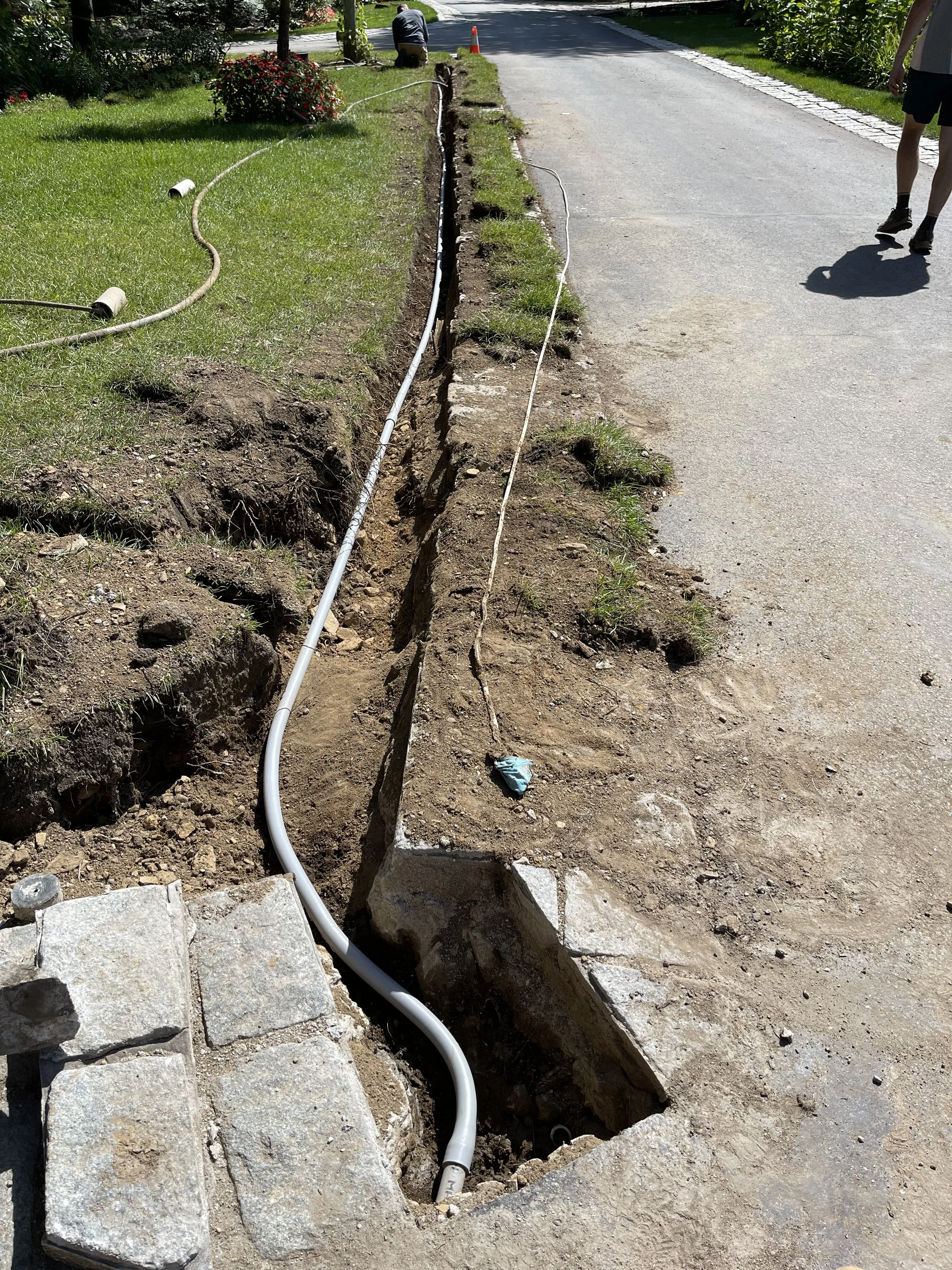 A sidewalk is under construction with a trench dug along the edge. White pipes are laid in the trench, and construction workers are present, with one kneeling in the background. The scene takes place next to a grassy area with red flowers and trees.