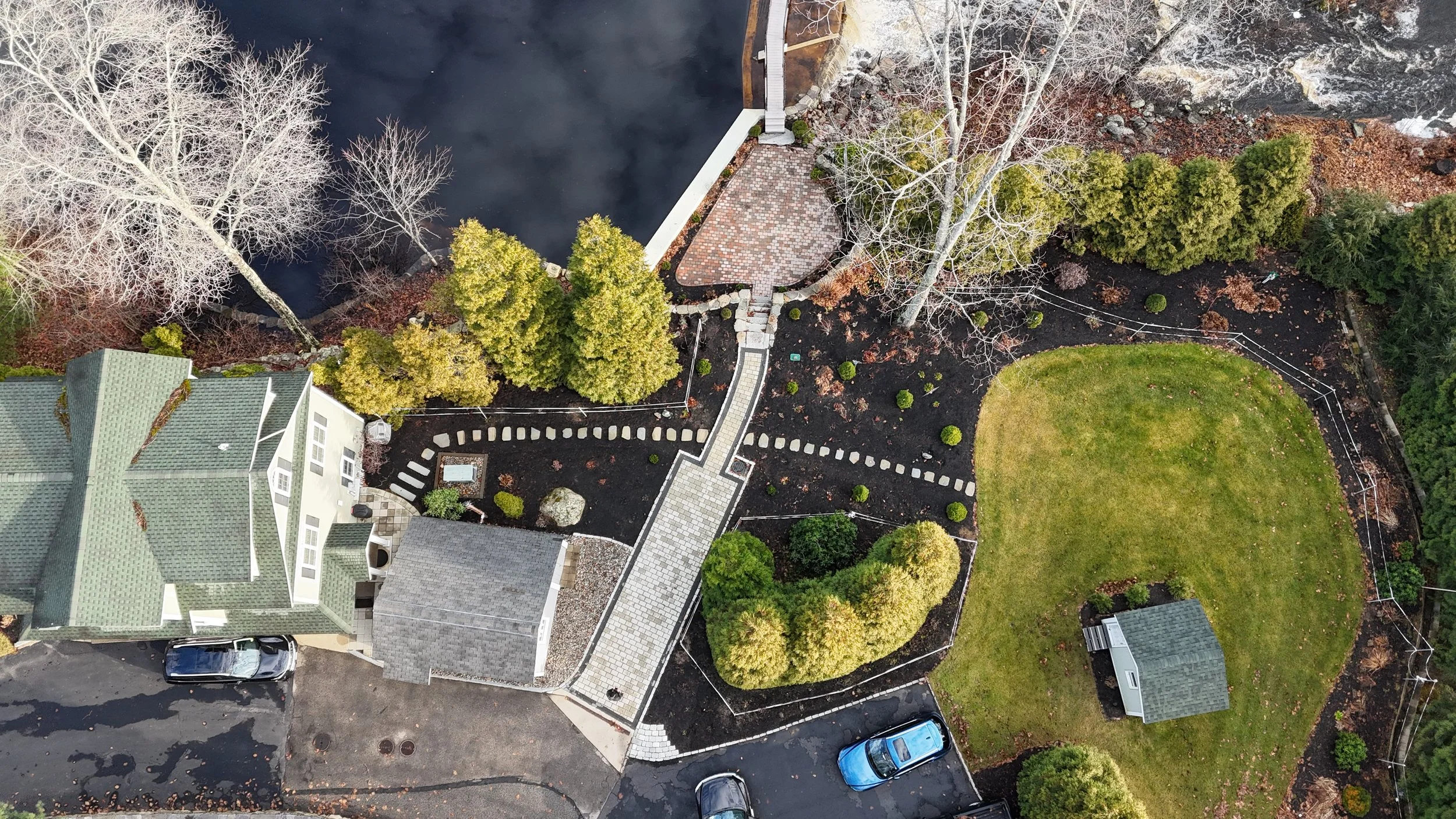 Aerial view of a residential yard with a stone pathway, a patio, trees, and green grass, surrounded by a driveway with parked cars.