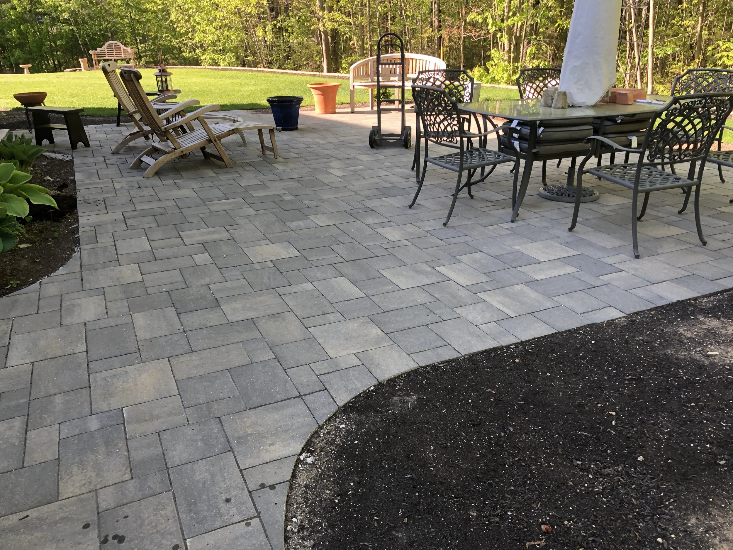 A backyard patio with a stone paver surface, outdoor furniture including a glass-top table and chairs, wooden lounge chairs, potted plants, and a grassy lawn with trees in the background.