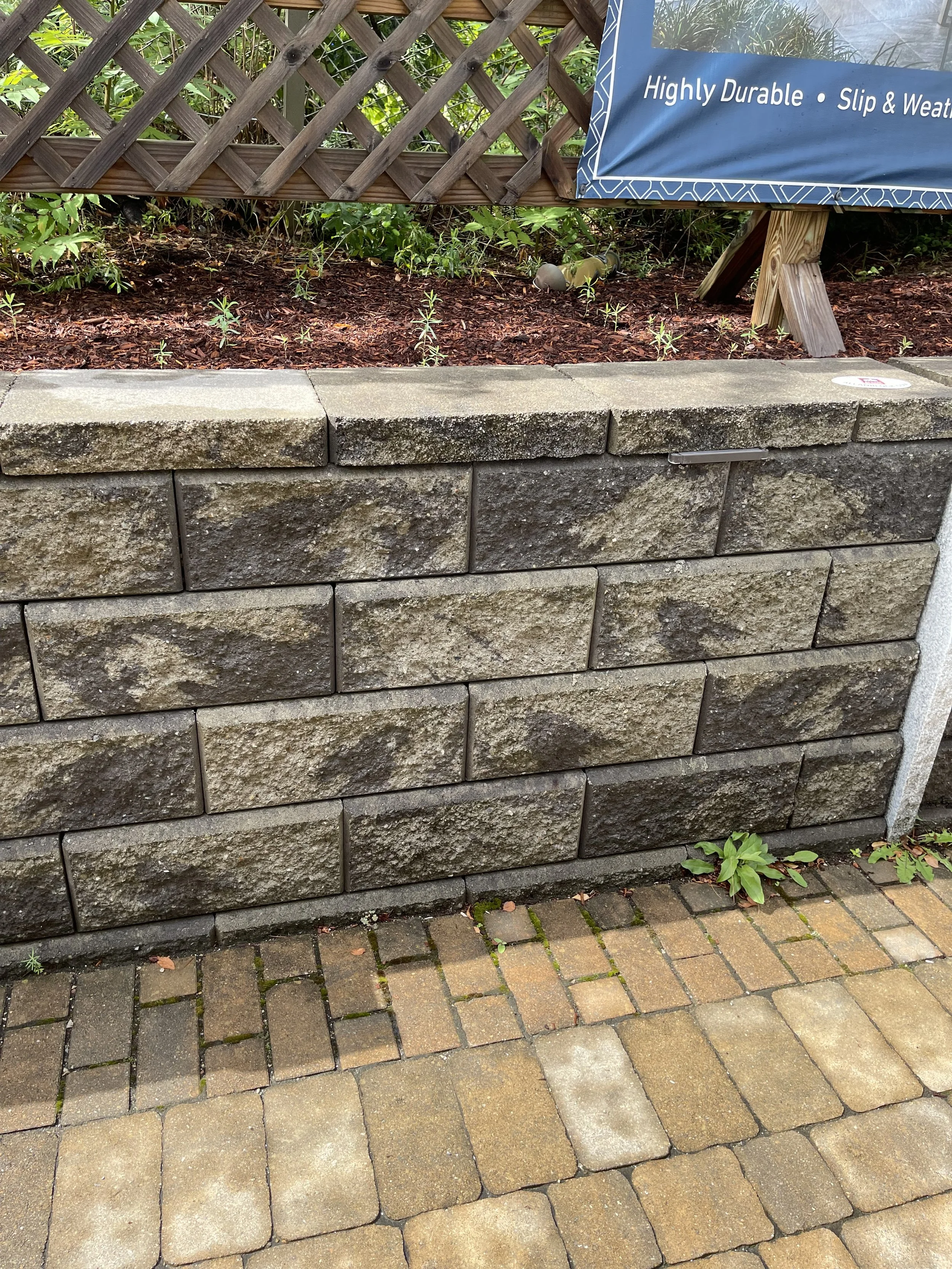 A low stone retaining wall with a gardening bed above it. The bed has some small plants and mulch, a lattice fence, and a blue banner. The ground in front is paved with bricks and concrete paving stones, with some greenery growing between the stones.