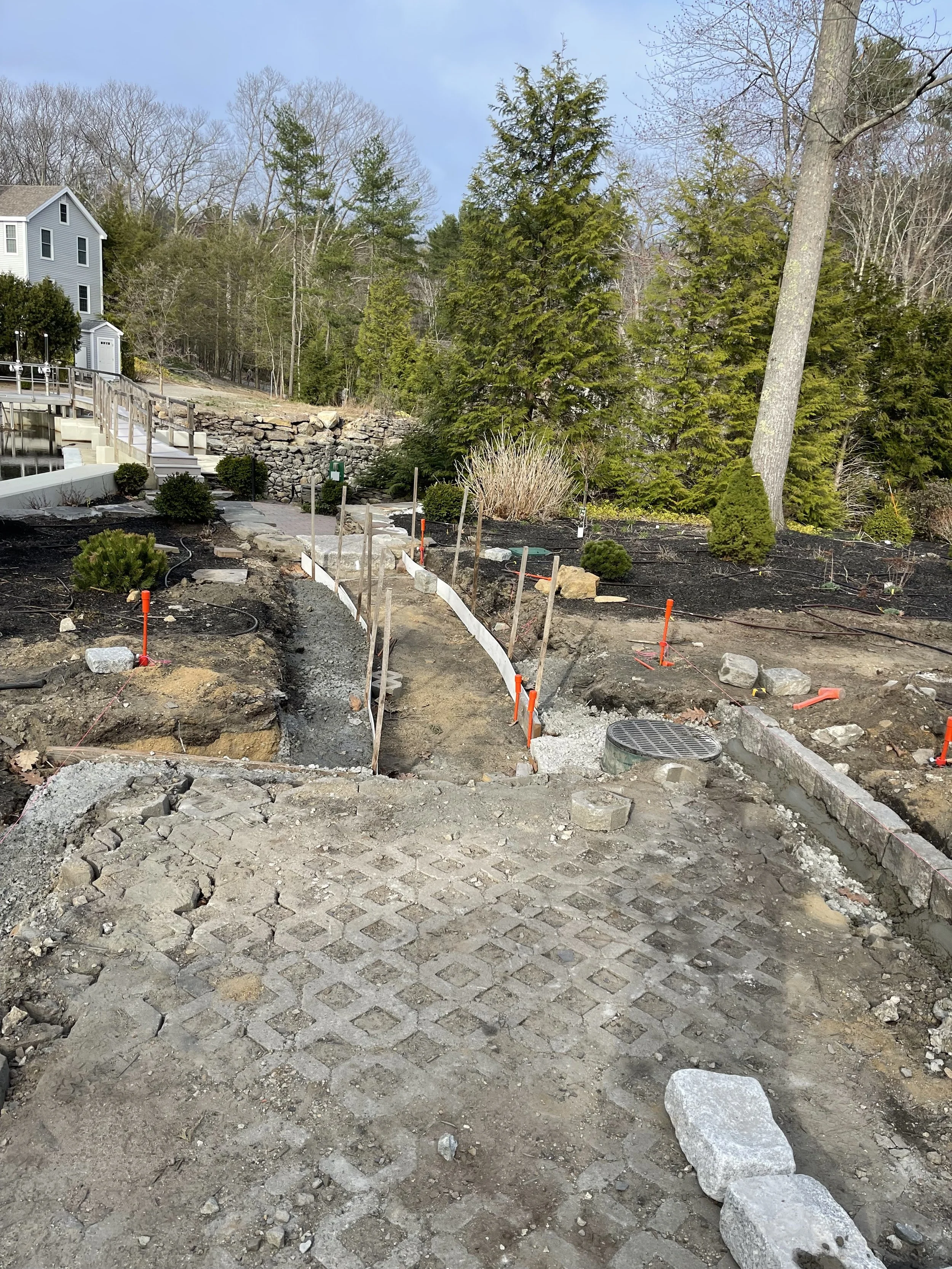 Construction site with a partially paved pathway, orange markers, and rocks on the ground, surrounded by trees and nearby houses.