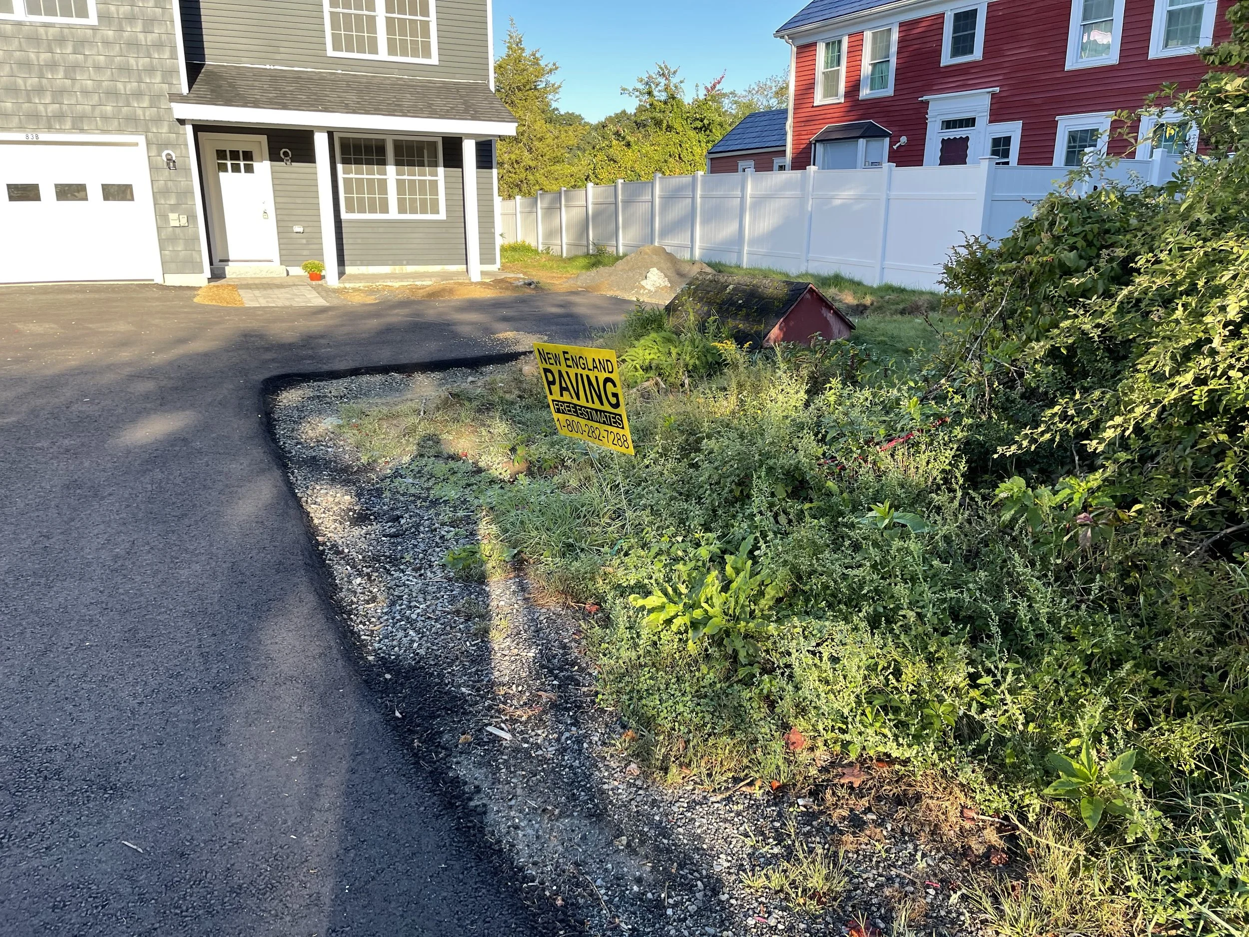 A residential backyard with a construction site for paving, a pile of dirt, and a sign that reads "New England Paving Free Estimates 1-800-282-7288." There are two houses in the background, one gray and one red, separated by a white fence. Green bush