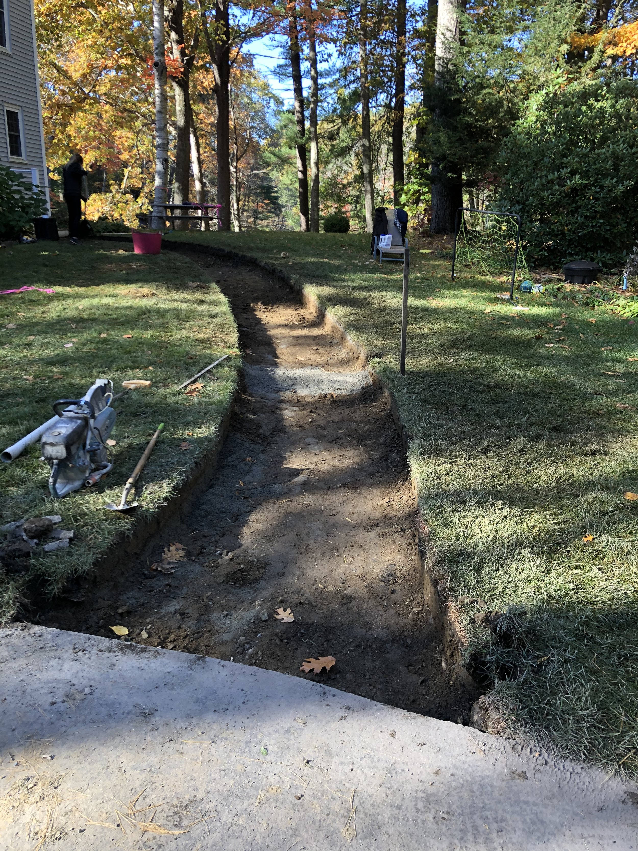 A garden area with a trench being dug for a new pathway, surrounded by trees and bushes with fall foliage, tools and construction materials nearby.