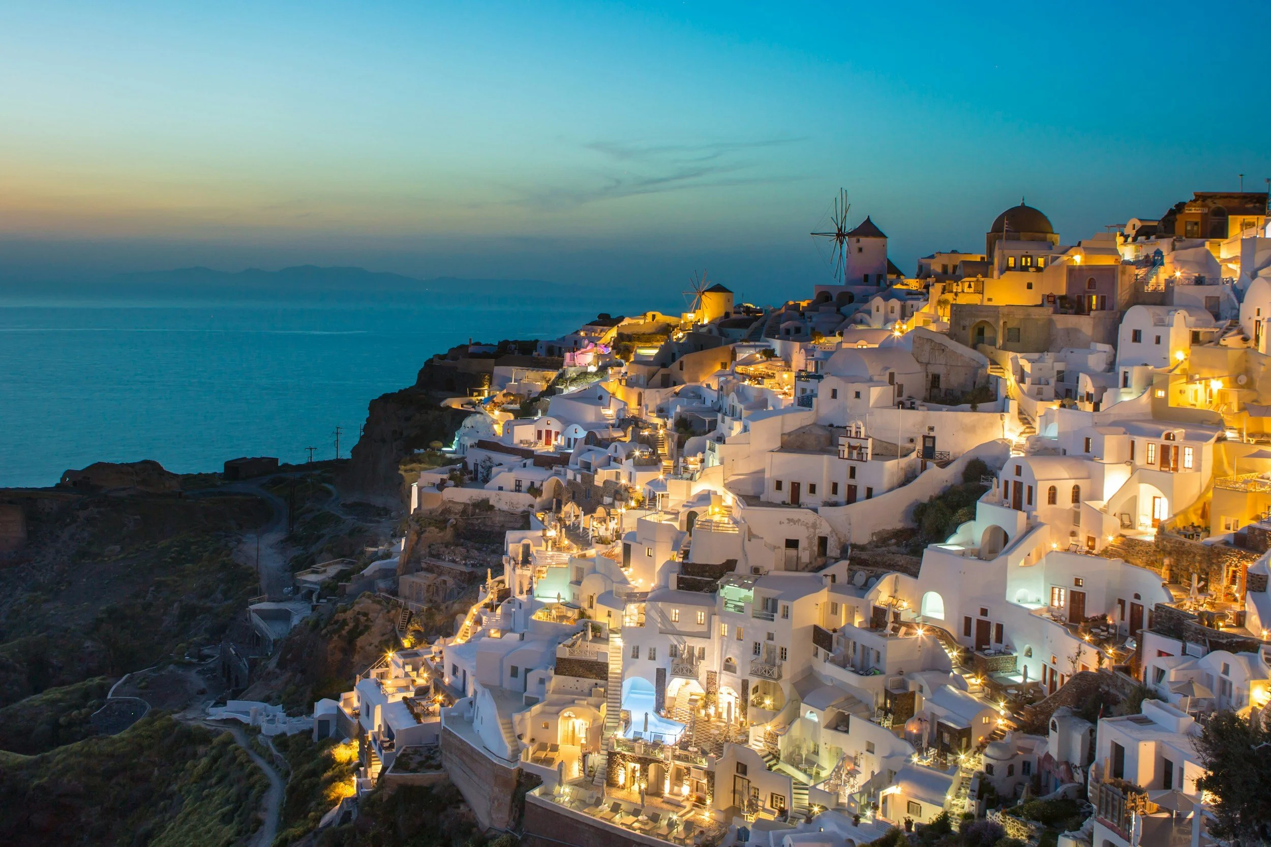 Santorini Greece at sunset with illuminated white buildings and windmill overlooking the Aegean Sea.