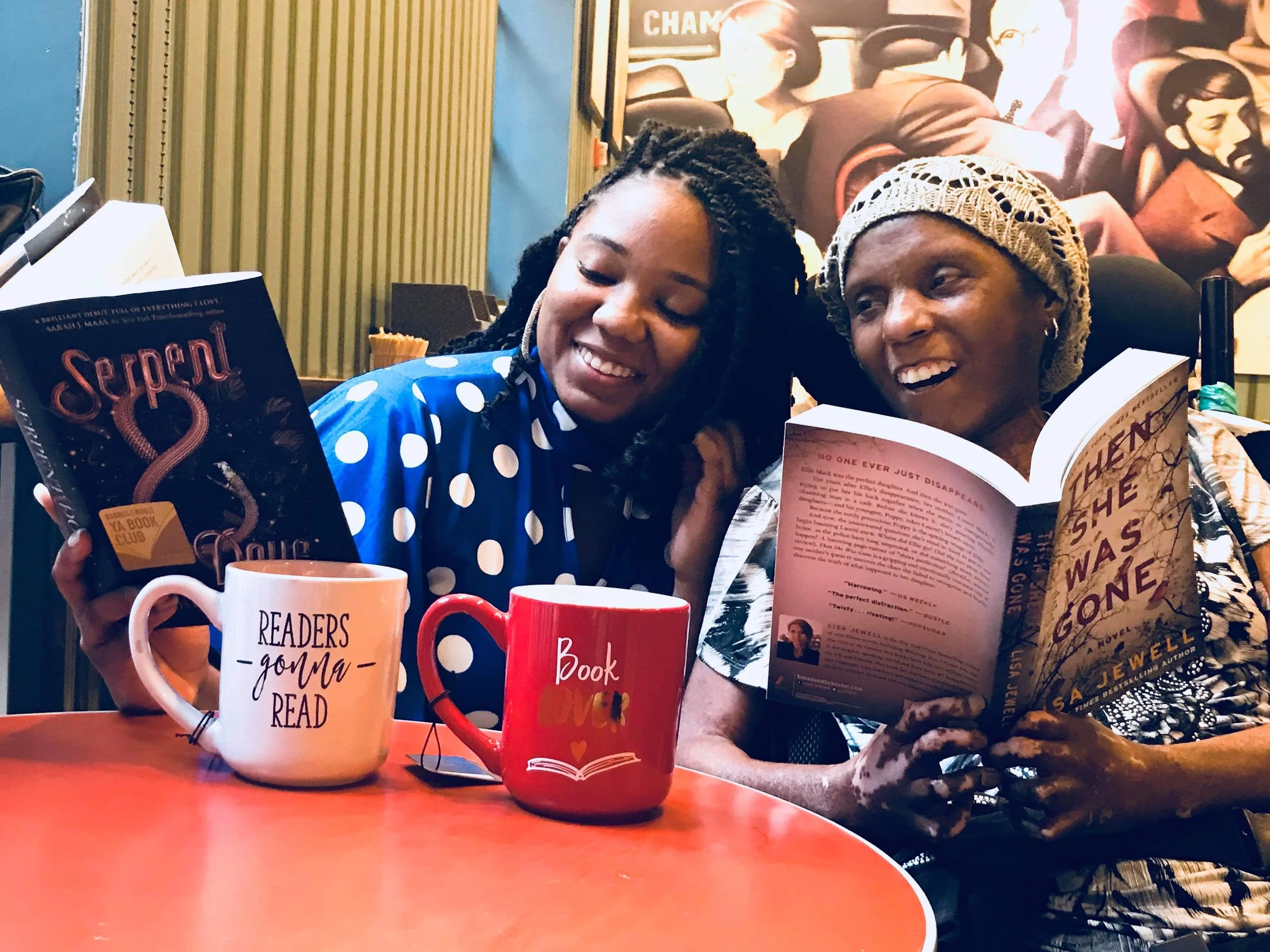 Two women sitting together at a table, reading books. The woman on the left has braided hair and is wearing a blue polka dot shirt, holding a book titled 'Serpent'. The woman on the right has a crochet hat and earrings, holding a book open in her lap. There are three mugs on the table with humorous and encouraging phrases, including 'Readers gon' read'. The background features a colorful mural with various figures.