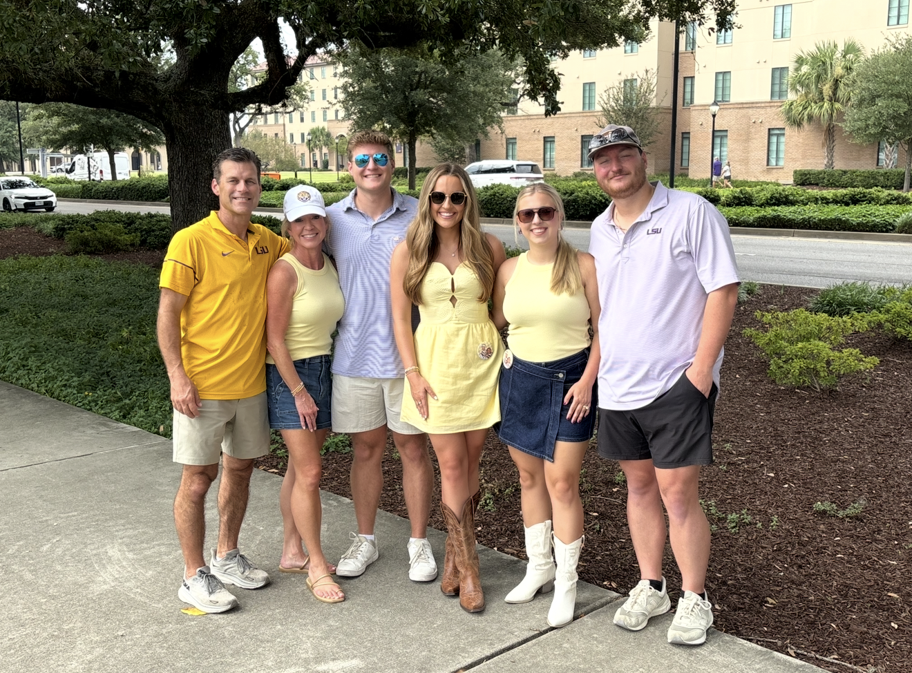 Group of six people standing outside in front of a large tree and building, smiling for a photo. They are dressed casually, with some wearing LSU clothing and sunglasses.
