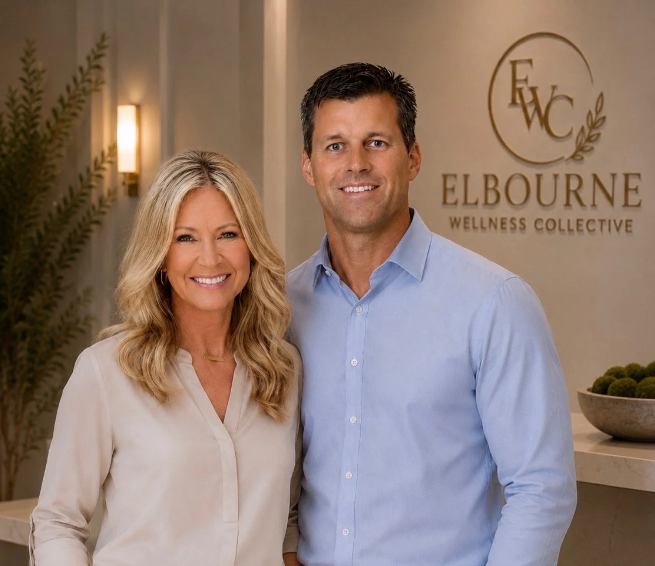 A smiling woman and man standing inside a wellness center reception area with Elbourne Wellness Collective sign in the background.