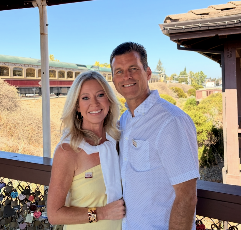 A smiling man and woman standing together outdoors on a sunny day, with a mountain train in the background and a railing decorated with padlocks.