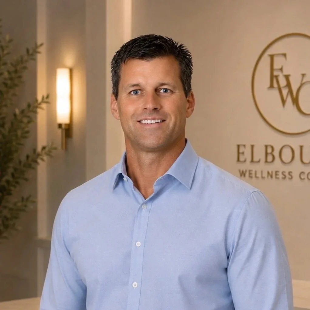 A man with dark hair wearing a light blue dress shirt smiling at the camera, standing in front of a beige wall with a logo that says "Elbow Wellness Center".
