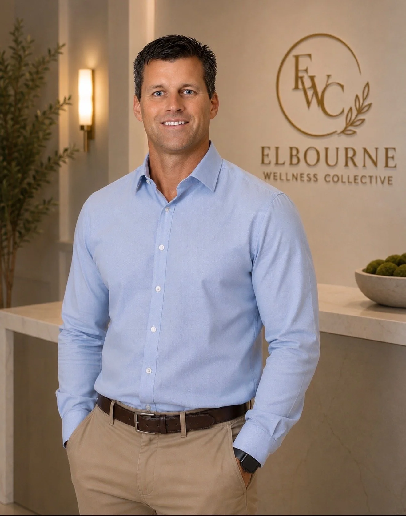 Man in light blue dress shirt and khaki pants standing in wellness center lobby, with 'Elbourne Wellness Collective' sign in the background.
