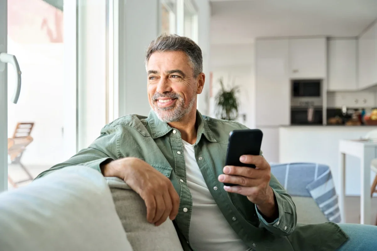 A middle-aged man with gray hair and beard sitting on a couch, smiling and holding a smartphone in his right hand, in a bright modern kitchen and living room.