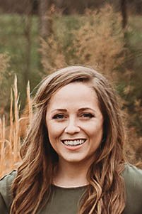 Lindsey Sims, Flourish Dietitian, with long, wavy hair standing outdoors in front of trees and tall grass.