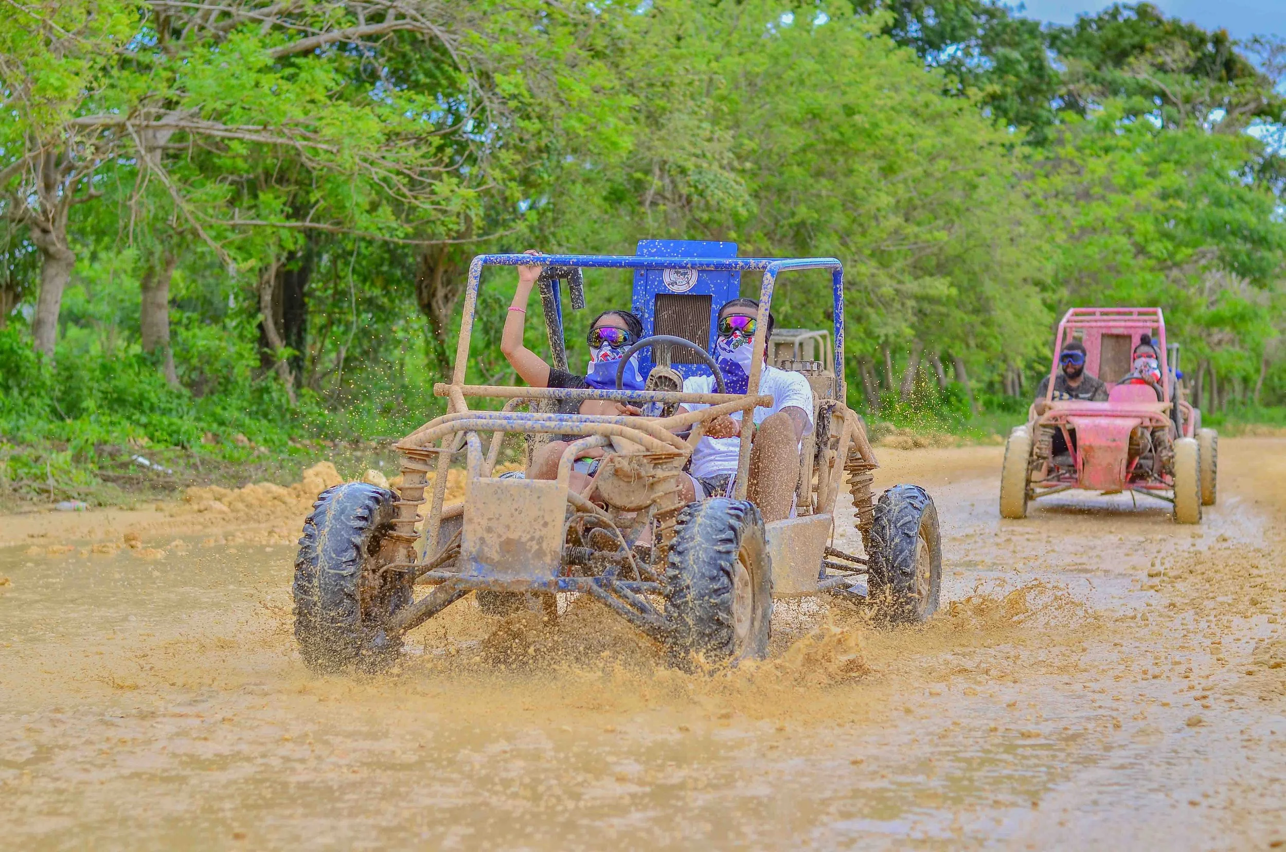 Two people driving off-road vehicles through a muddy terrain surrounded by green trees, with water splashing around the tires.