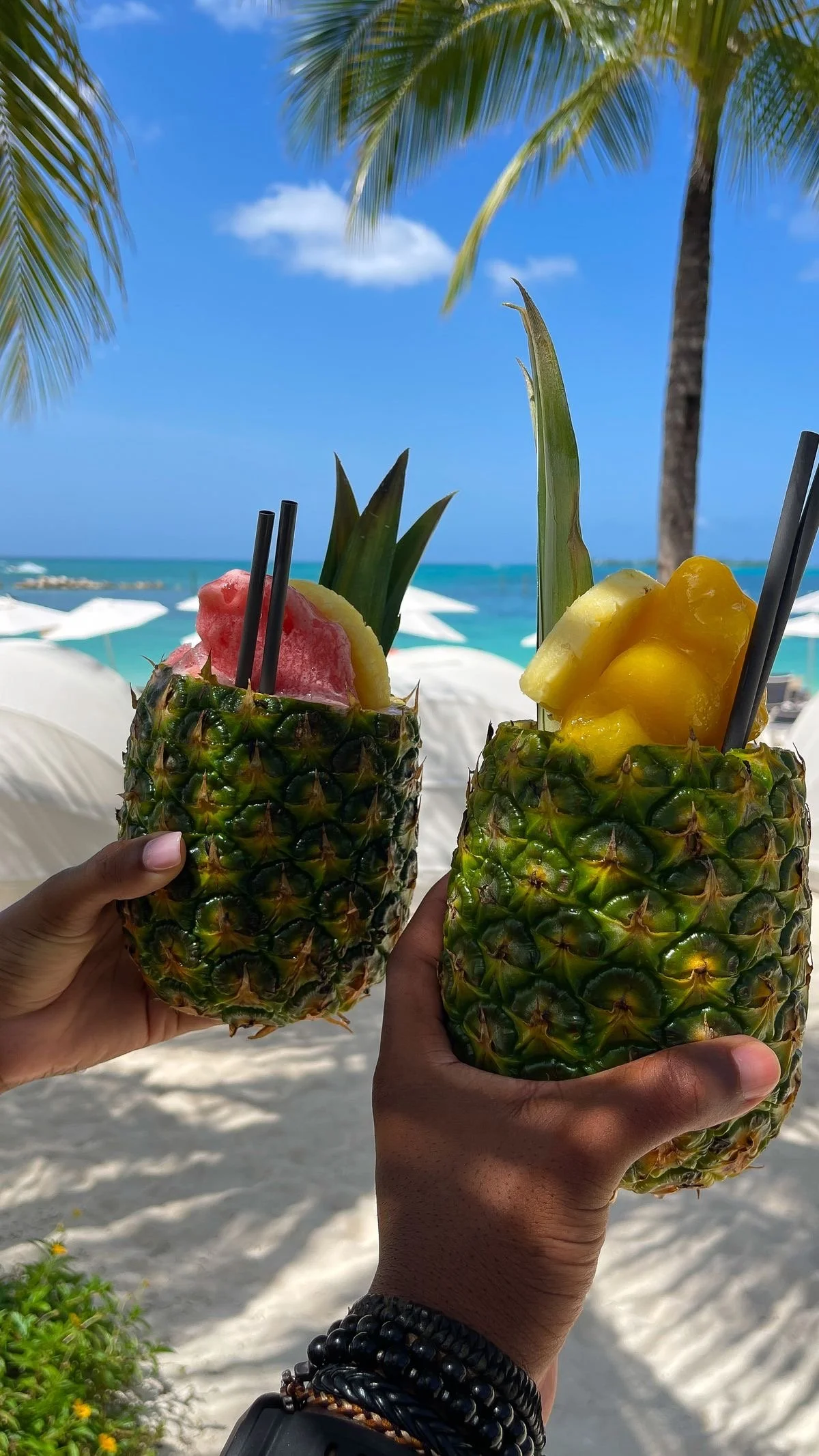 Two pineapples with their tops cut out, converted into drinks with colorful fruit and ice inside, being held up in front of a beach scene with blue sky, ocean, palm trees, and white umbrellas.