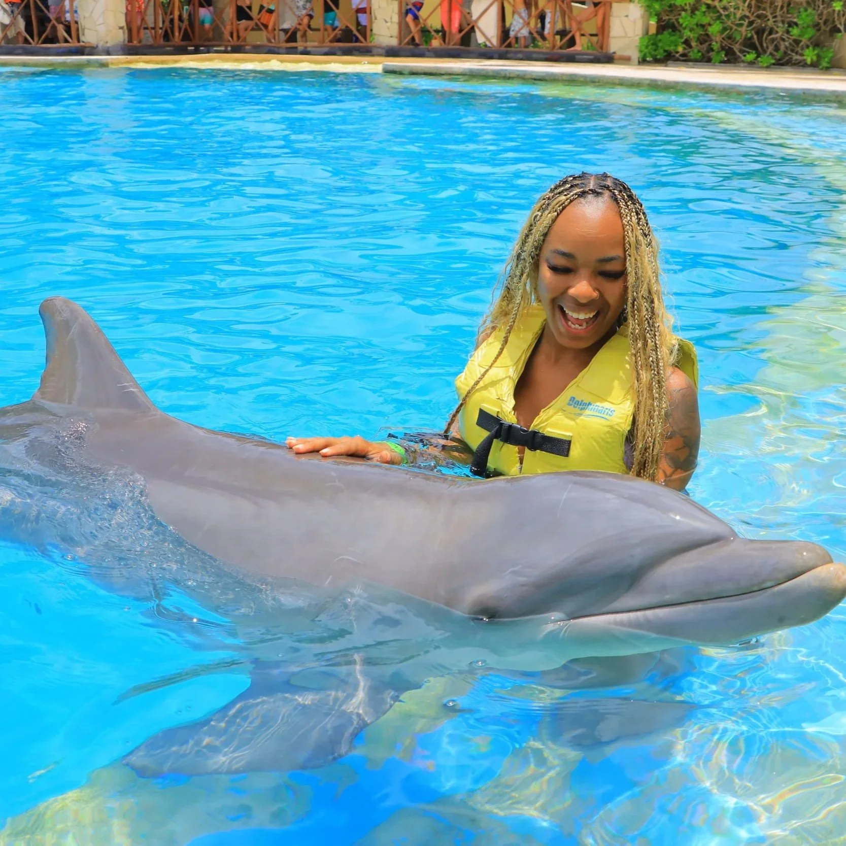 A woman with long blonde braided hair in a yellow life jacket standing in a swimming pool, smiling while holding onto a dolphin's dorsal fin partially submerged in water, with a background of people on a wooden deck.
