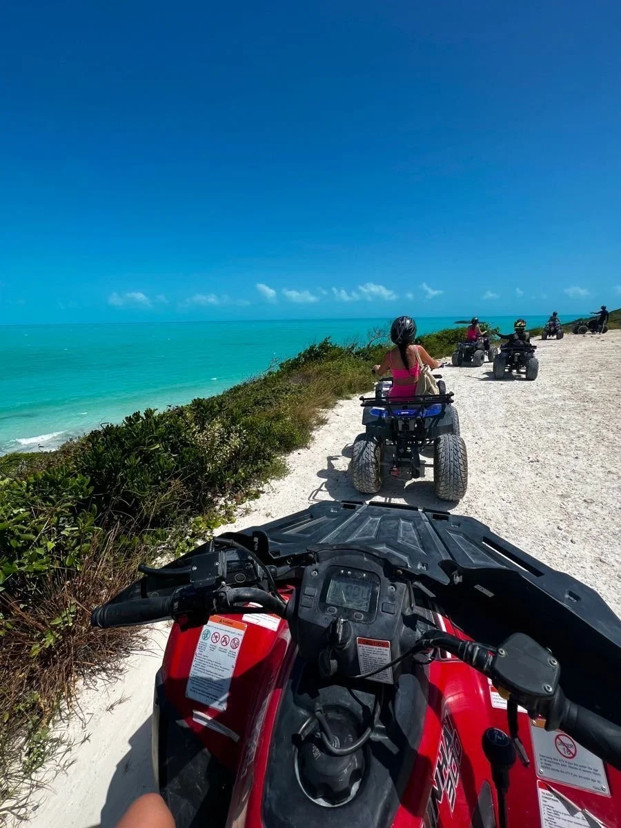 A group of people riding all-terrain vehicles (ATVs) along a coastal trail with white sand and green shrubbery, ocean and blue sky in the background.