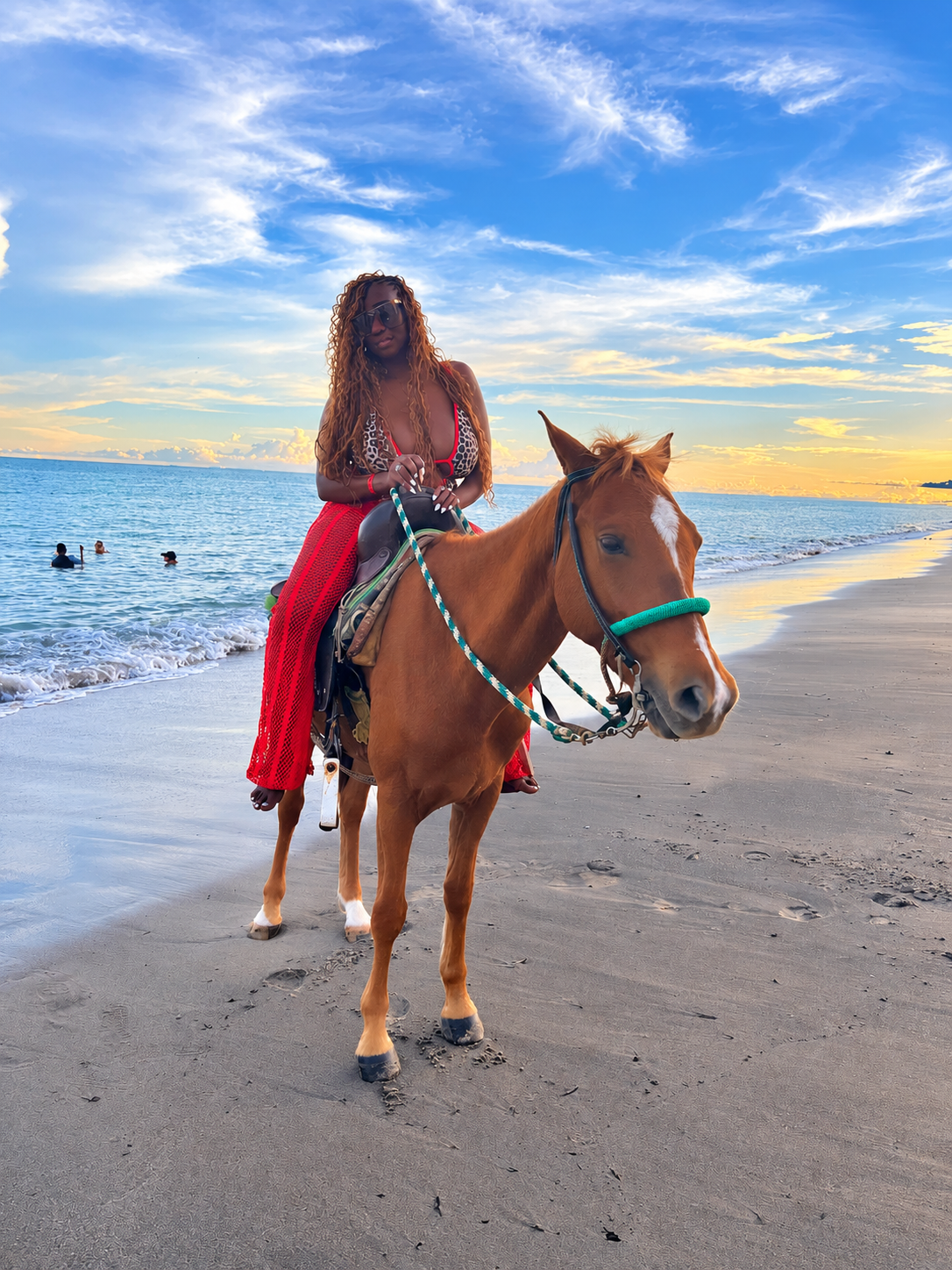 A woman riding a horse on a beach at sunset, with beachgoers in the water and a colorful sky overhead.